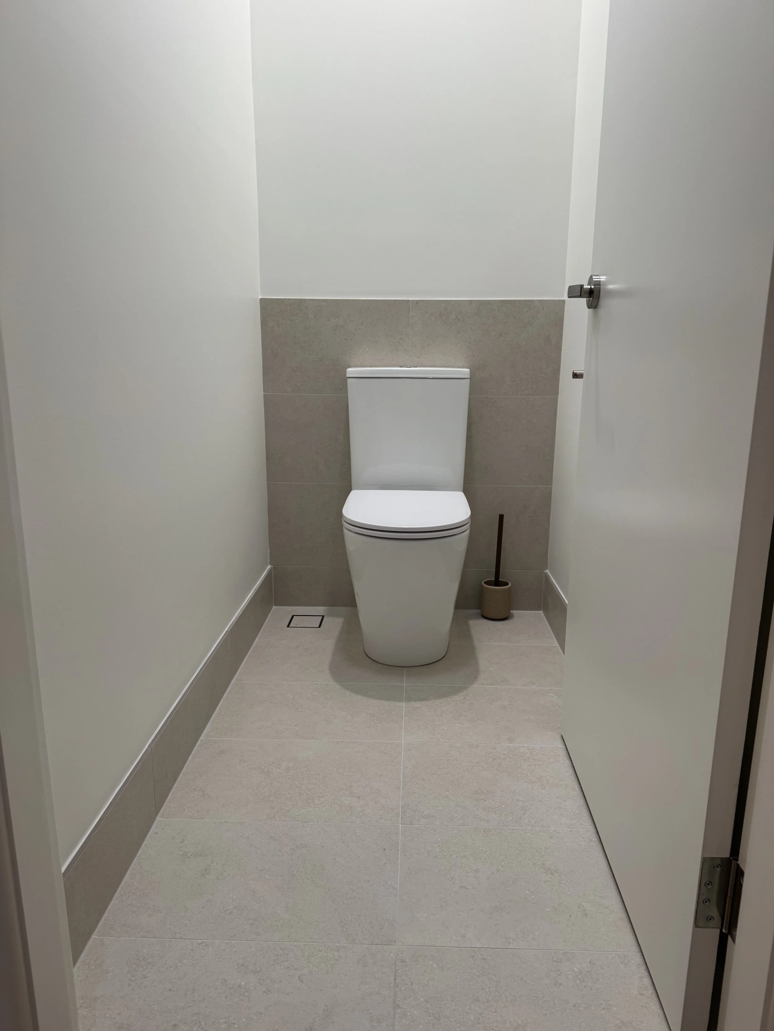 A clean, modern bathroom with a white toilet, beige tiled wall behind it, and a small toilet brush in a holder on the floor. The bathroom has light-colored floor tiles and white walls.