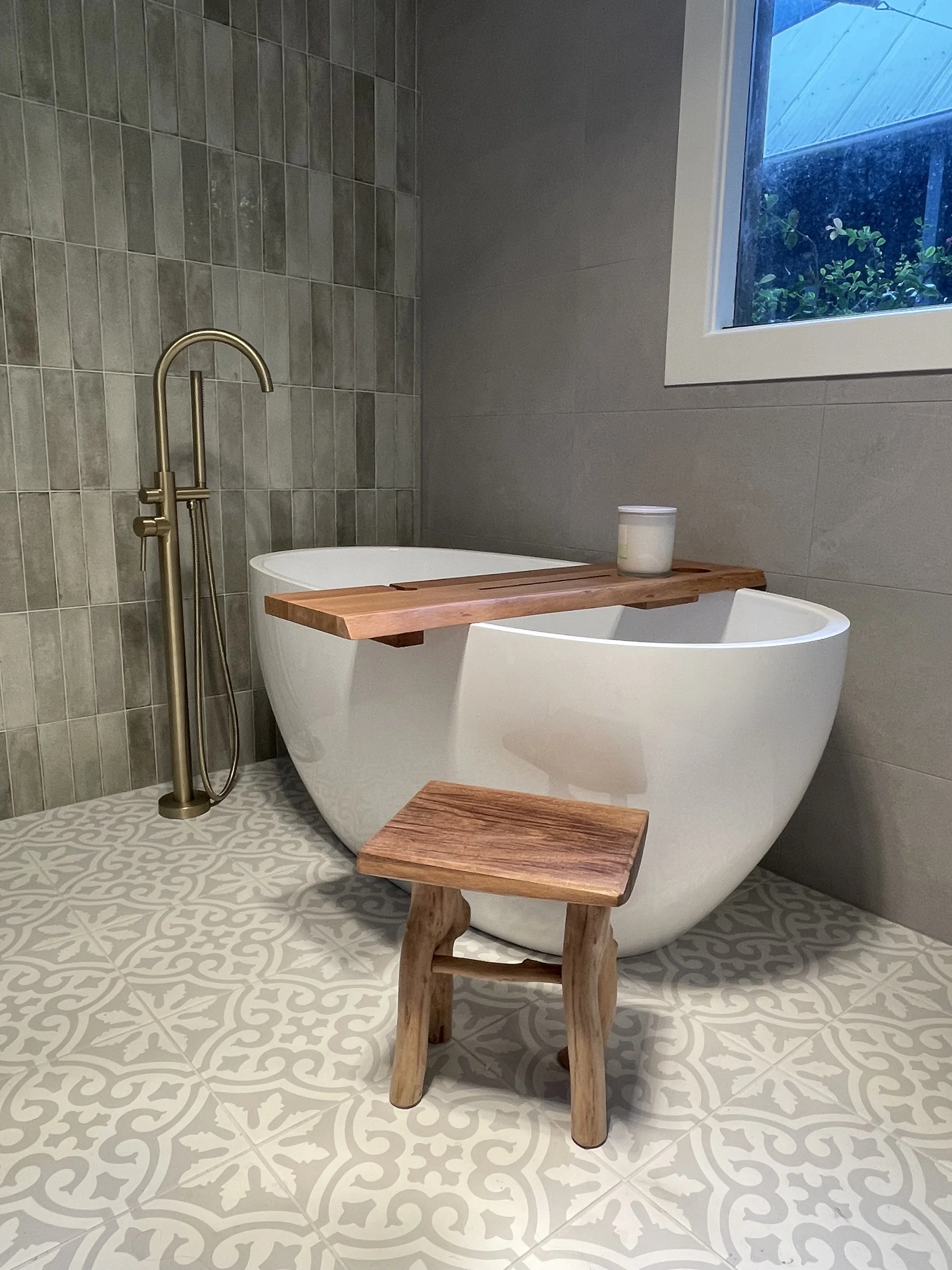 A modern bathroom with gray tiled walls and patterned floor tiles. There is a white freestanding bathtub with a wooden tray and a small wooden stool. A brass floor-mounted bathtub faucet is next to the tub. A window shows greenery outside.