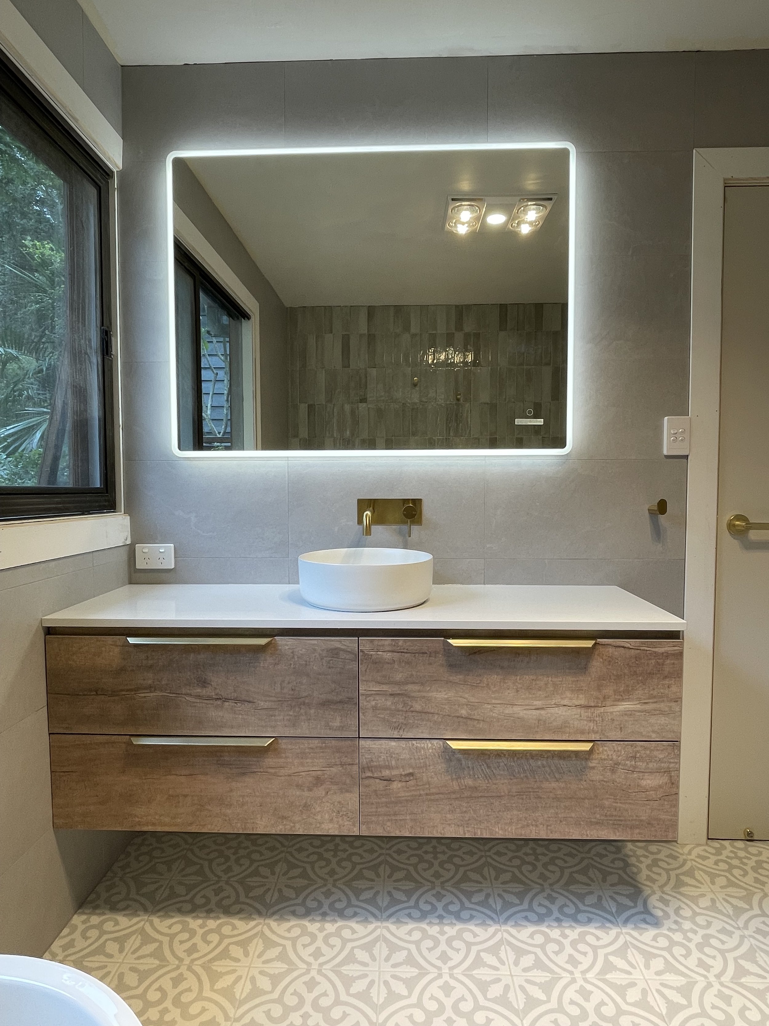Modern bathroom vanity with a white vessel sink, a large backlit mirror, and a wooden cabinet below. The wall has gray tiles, and there is a window to the left with a view of greenery outside. The floor has patterned tiles.