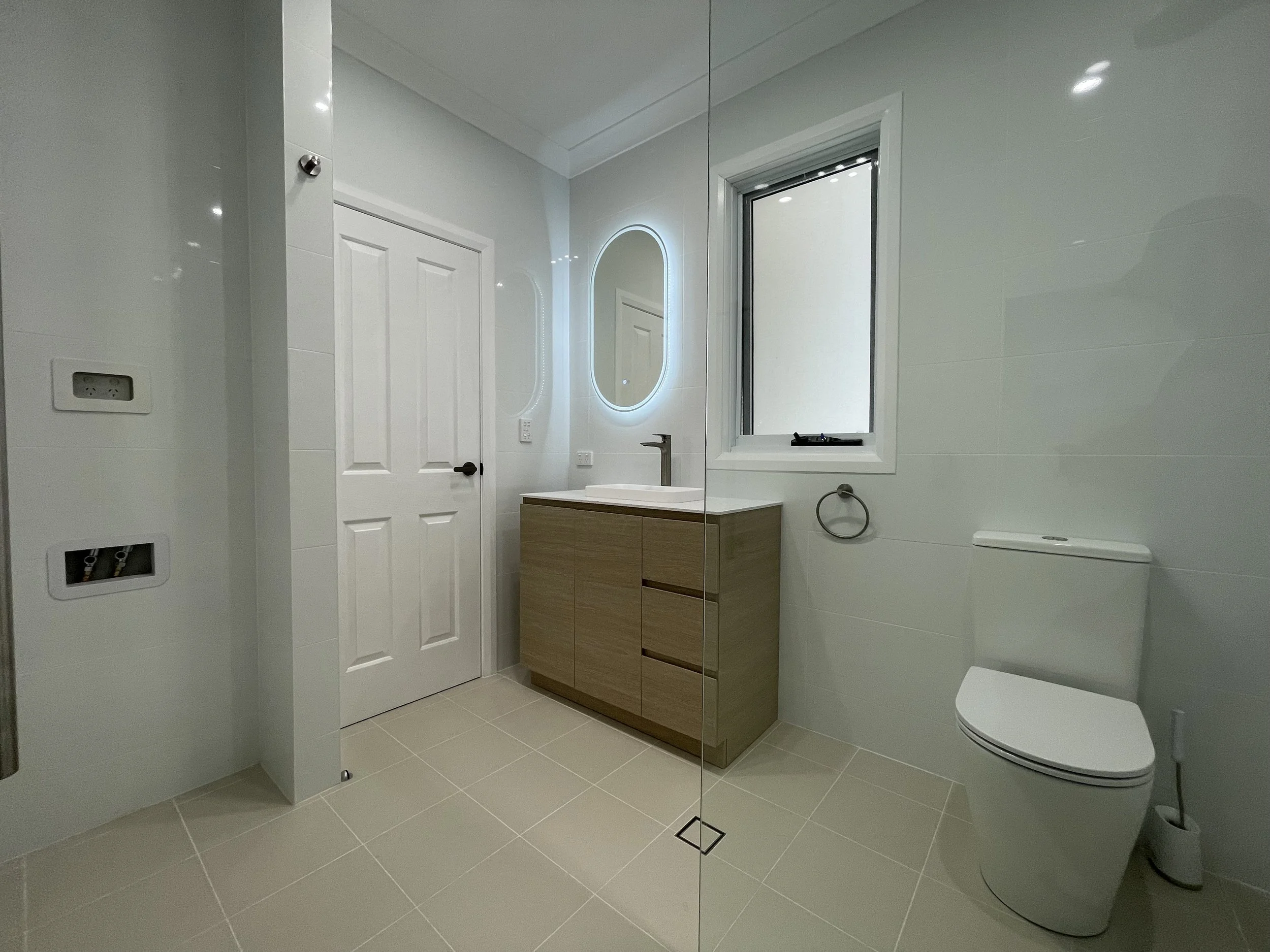 Modern minimalist bathroom with white walls, beige tile flooring, a wooden vanity with a white sink, a mirror with LED backlight, a window, and toilet.