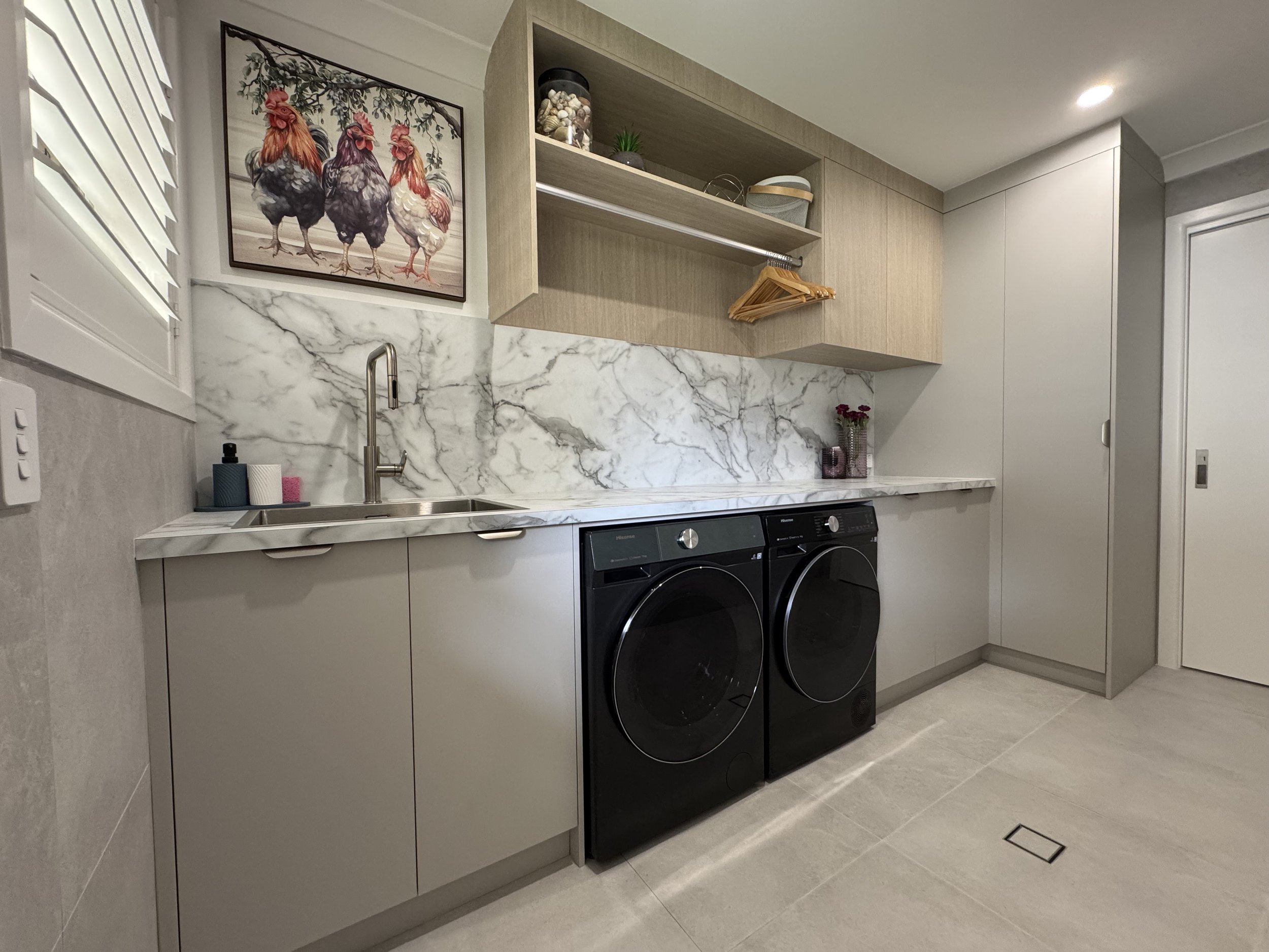 A laundry room with a marble countertop, beige cabinets, and a washer and dryer. There is a picture of chickens on the wall, plants, and a container of seashells on the shelf.