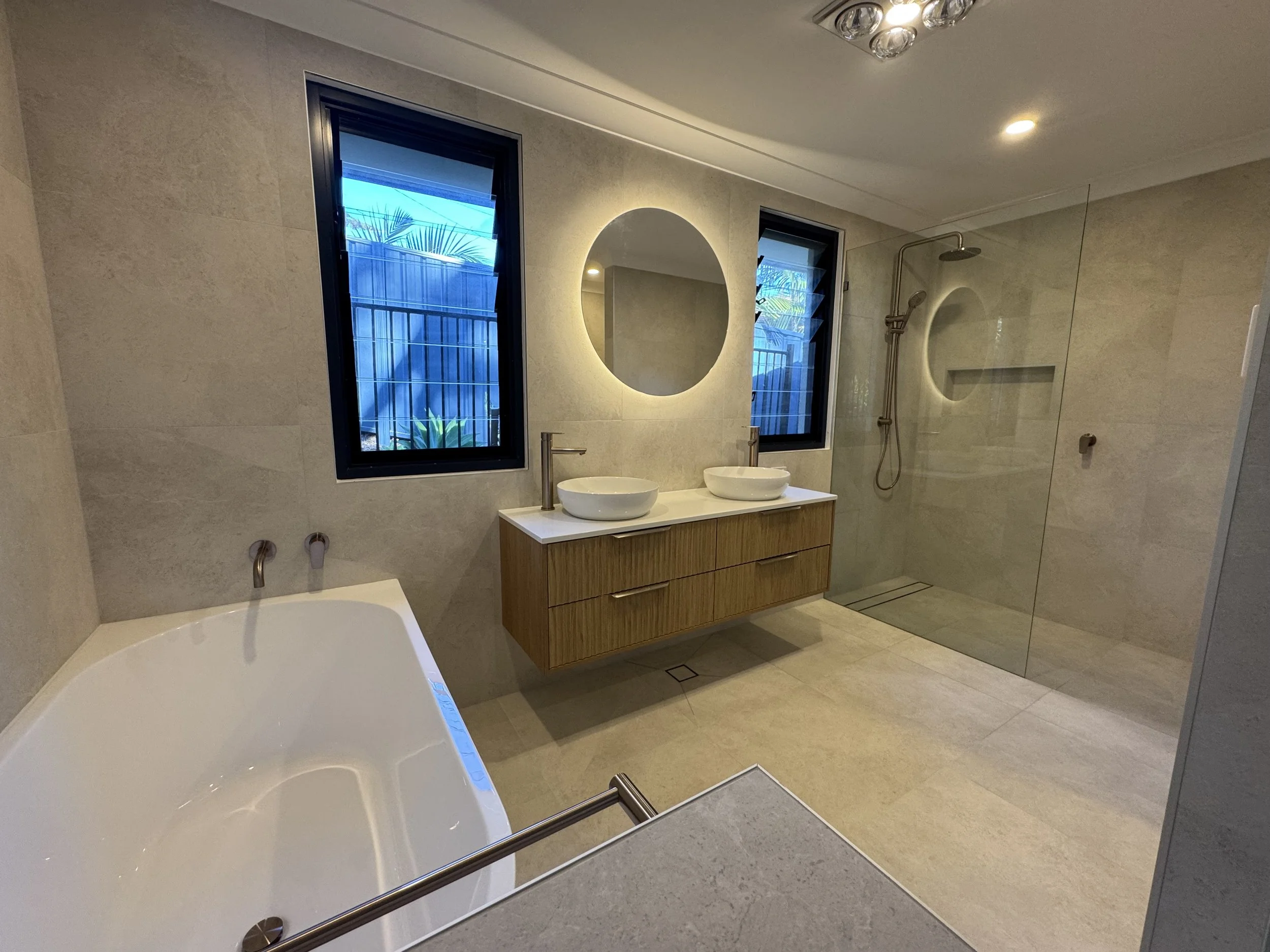 Modern bathroom with two oval vessel sinks on a wooden vanity, a large backlit round mirror, frosted glass windows, a walk-in shower with a rain shower head, a bathtub, and beige tiled walls and floor.