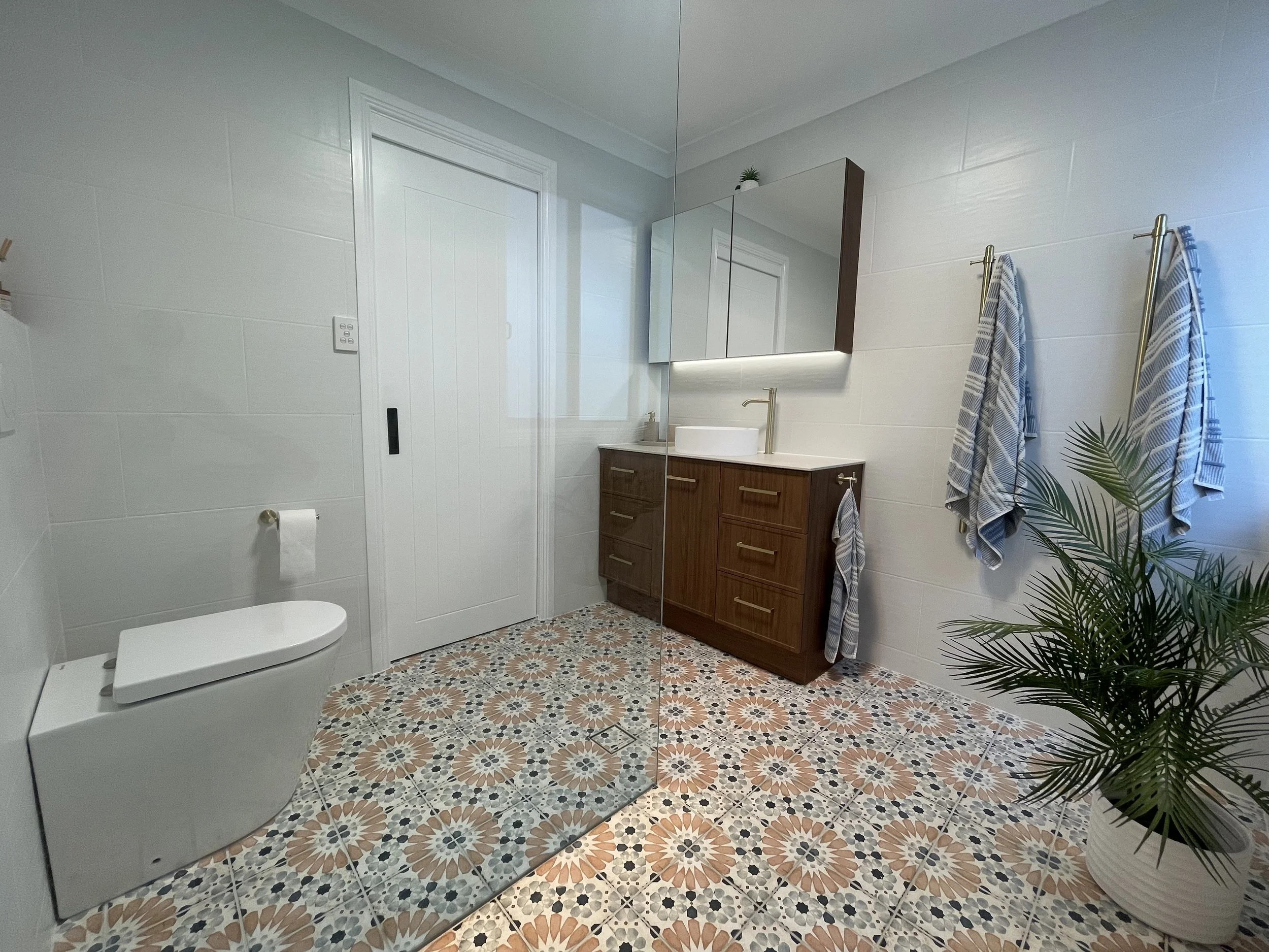 A bathroom with white tiled walls, a white modern toilet, a wooden vanity with a round vessel sink, a large mirror cabinet, and blue and white towels hanging on gold towel bars. There is a potted plant in the corner and patterned floor tiles.