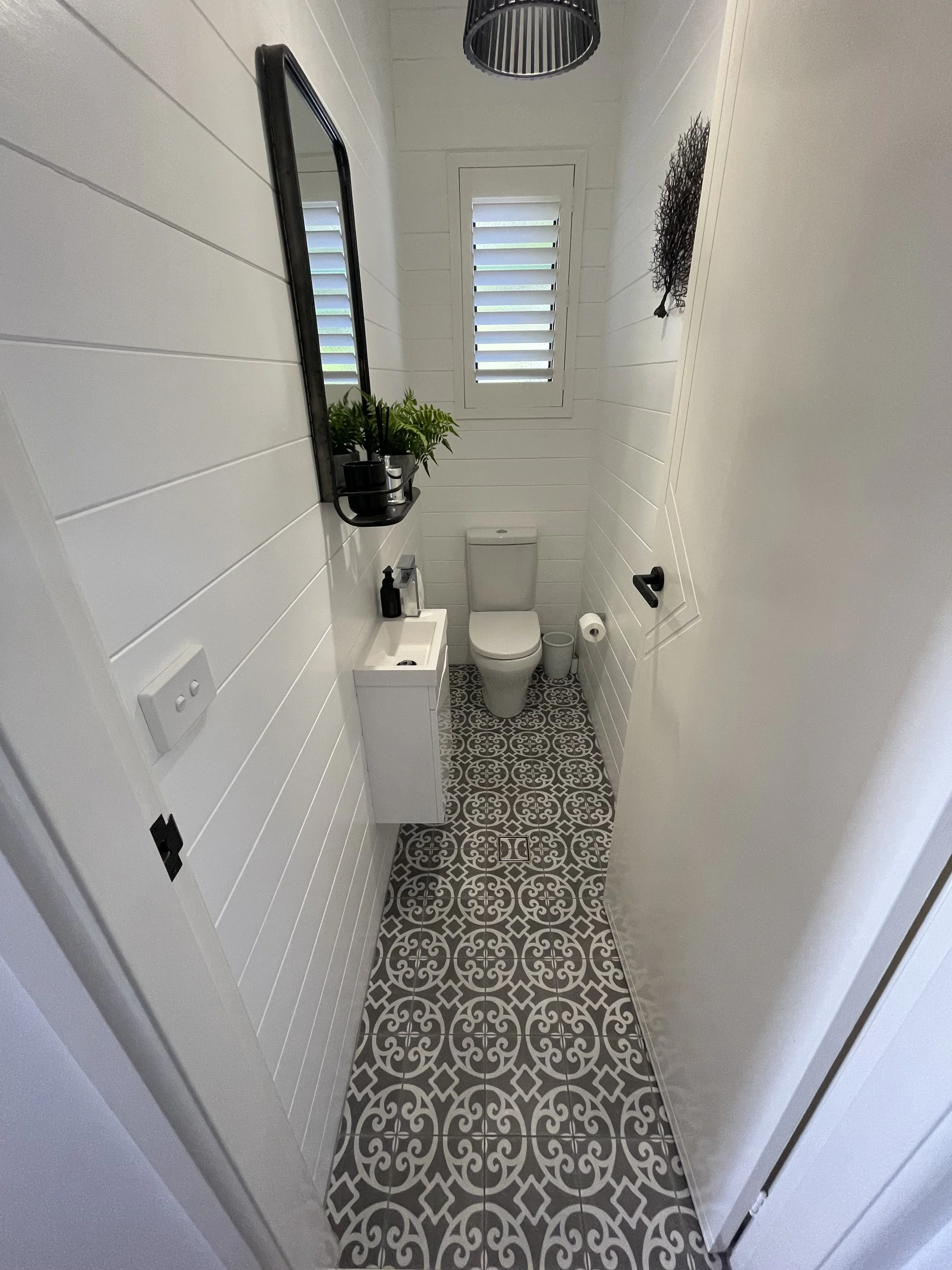 A small, narrow bathroom with white paneled walls, a small white sink, a toilet, and black fixtures. There is a window with horizontal blinds and patterned tile flooring. Decor includes a mirror, a small plant, and a wall art piece.