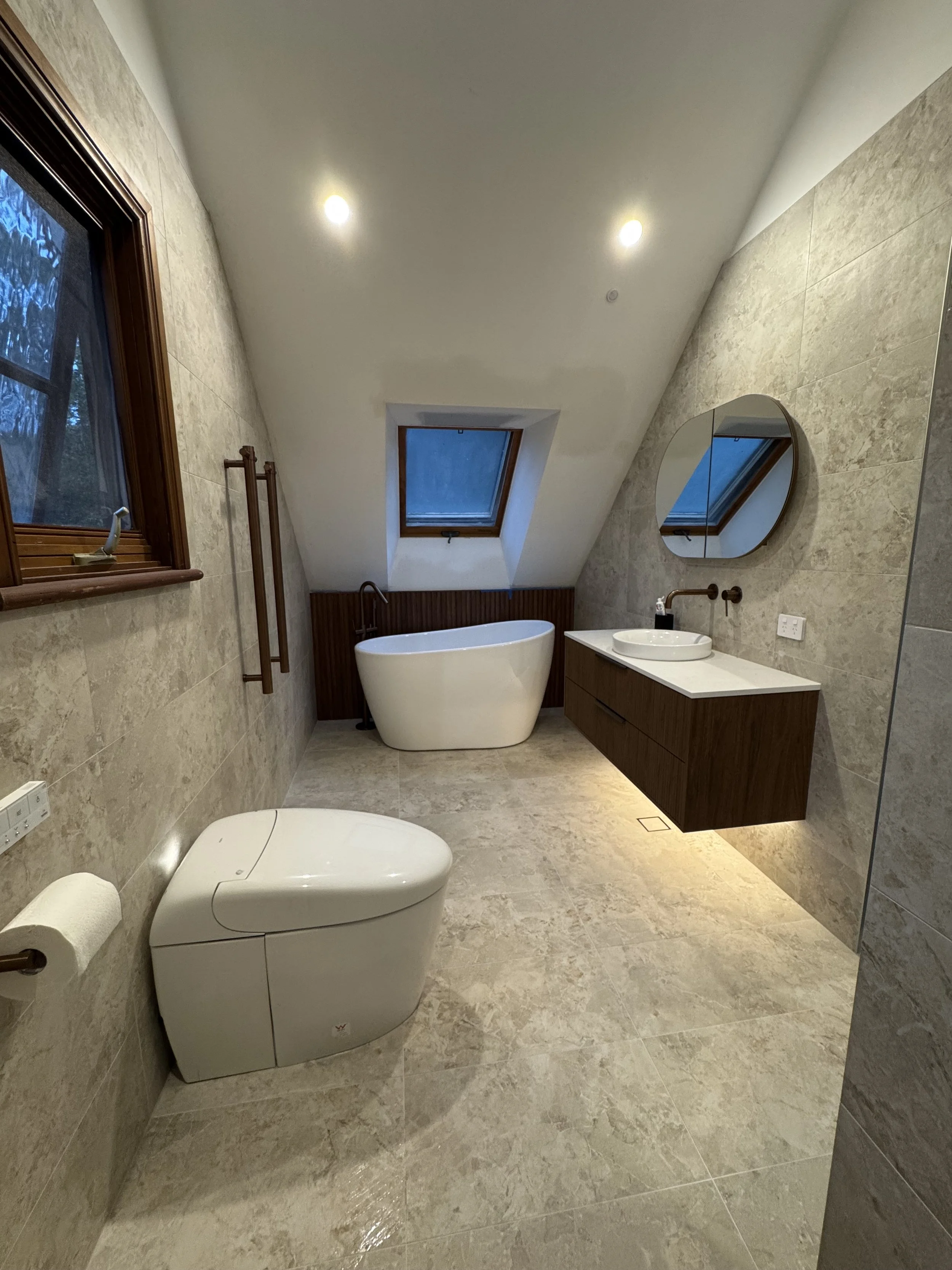 A modern bathroom with beige tiled floors and walls, a white freestanding bathtub under a skylight window, a wall-mounted vanity with a circular mirror, and a smart toilet with a paper holder on the wall.