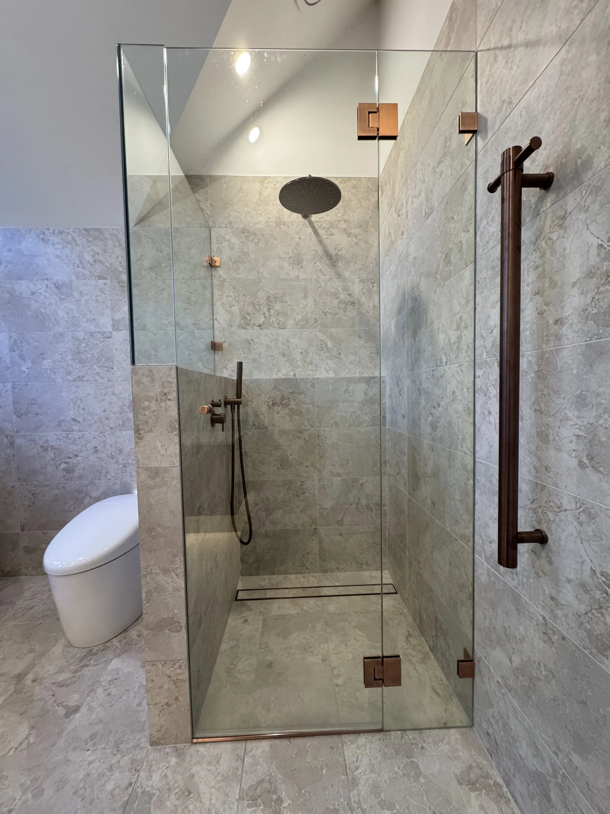 Modern bathroom with a walk-in shower enclosed by glass, featuring a rainfall showerhead. The bathroom has beige marble tiles on the walls and floor, with a white toilet visible to the left. A wooden grab bar is mounted on the right wall.