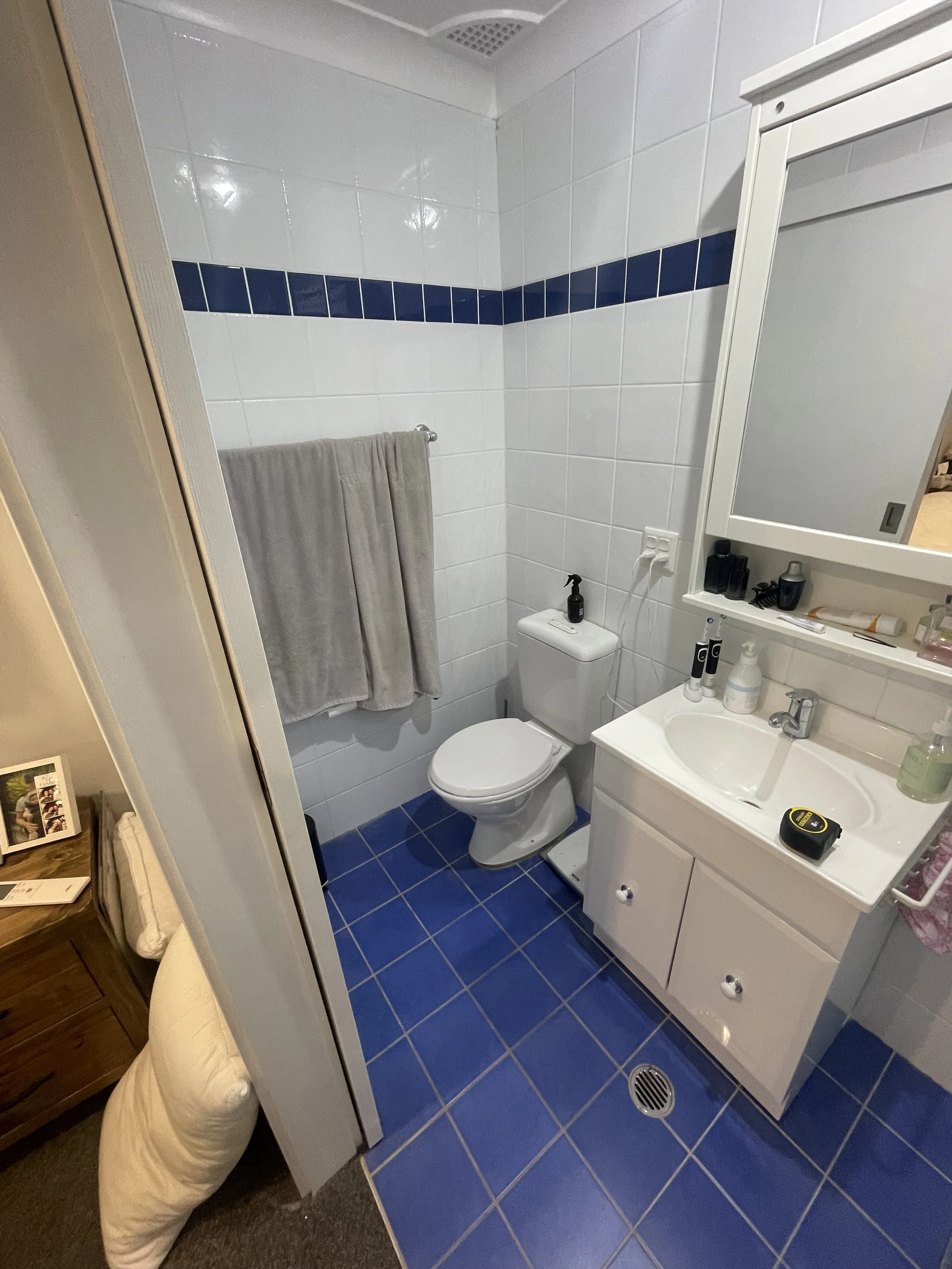 A small bathroom with white tiled walls, a blue tile border, a toilet, a sink with a mirror cabinet, and a towel hanging on a rail. There are various toiletries on the sink countertop and a small kabinet mirror.
