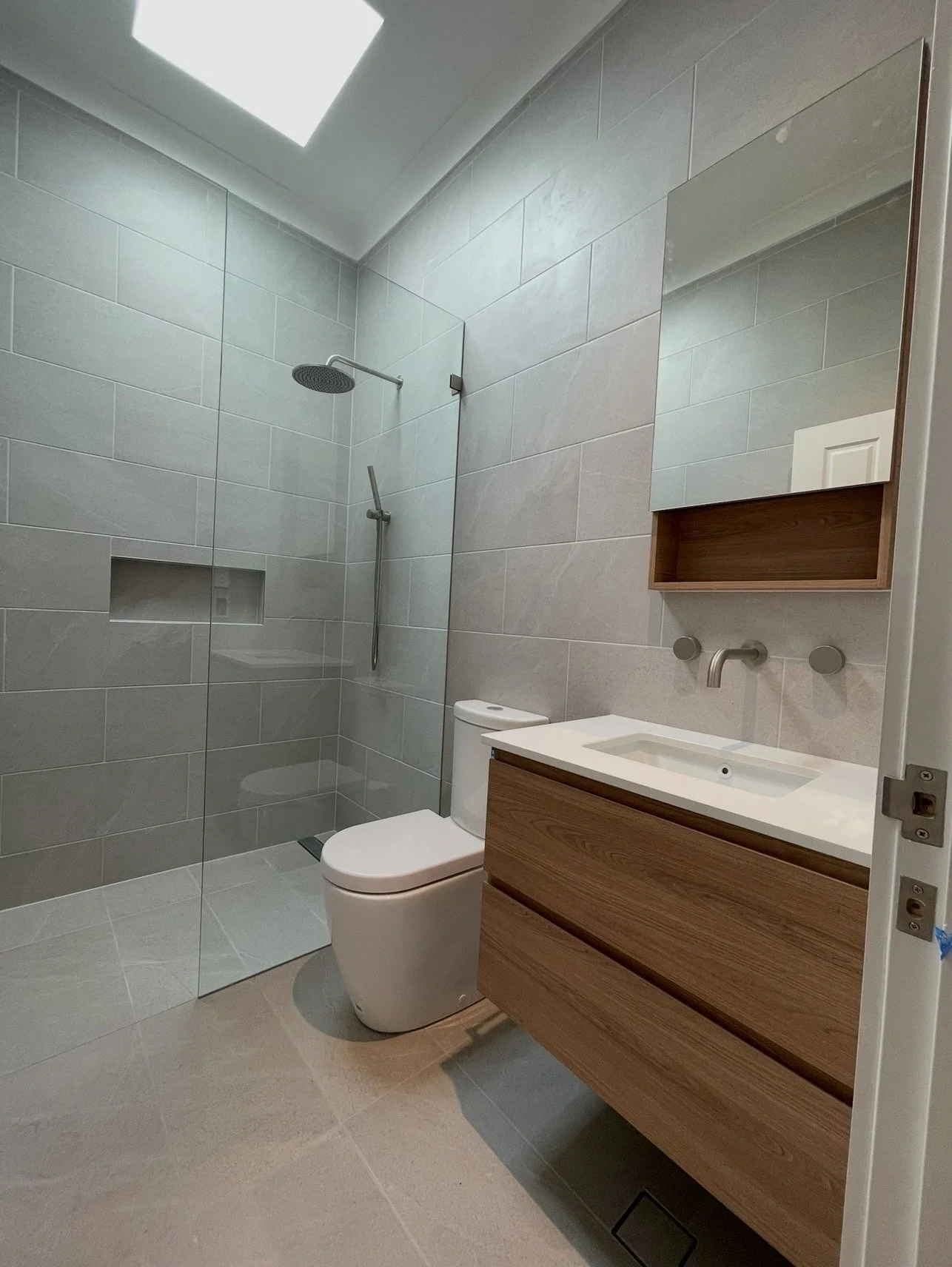Modern bathroom with gray tiled walls, glass shower enclosure, wall-mounted mirror, wooden vanity with sink, and a toilet.