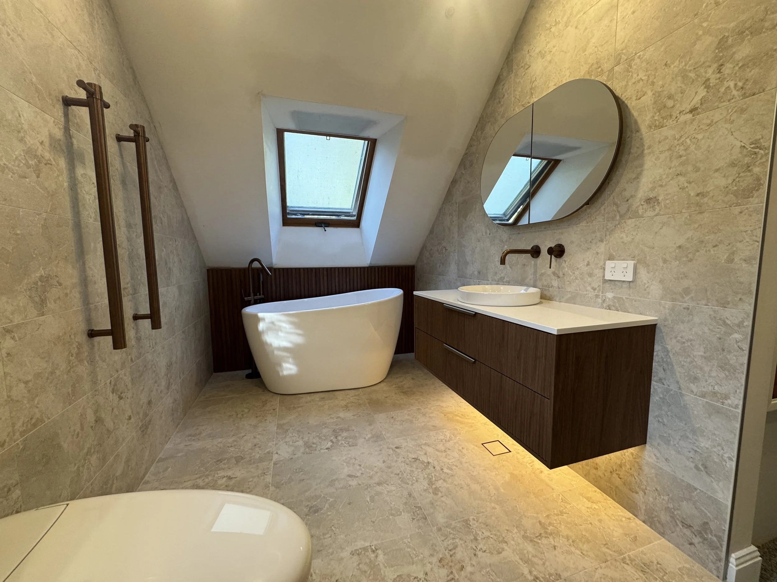 Modern bathroom with a sloped ceiling, skylight window, beige tiled walls and floor, white freestanding bathtub, round mirror above a wooden vanity, and a brass towel rack.