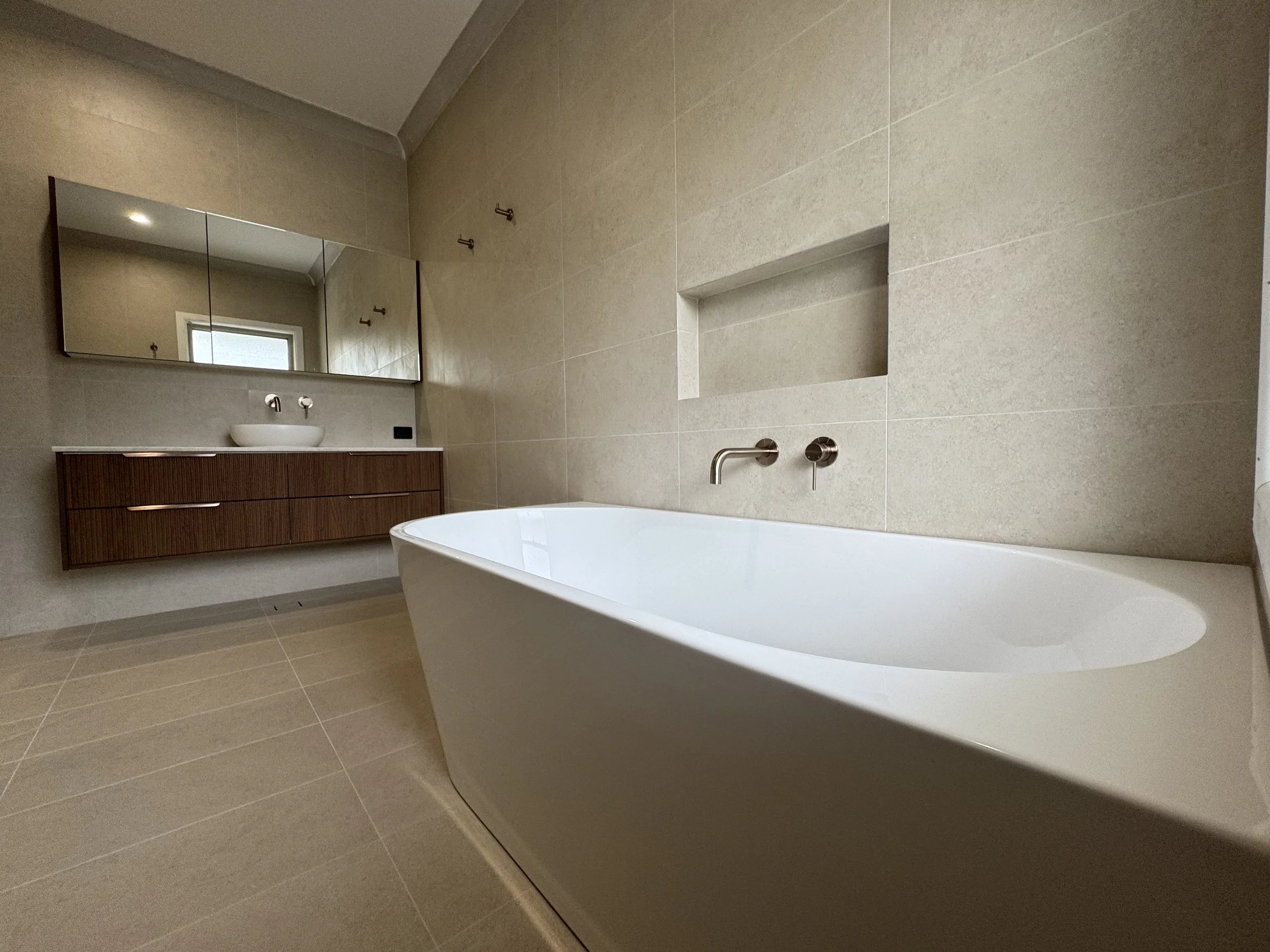 Modern bathroom with beige tiles, a freestanding bathtub, a large mirror, a wooden vanity with a vessel sink, and minimalistic fixtures.