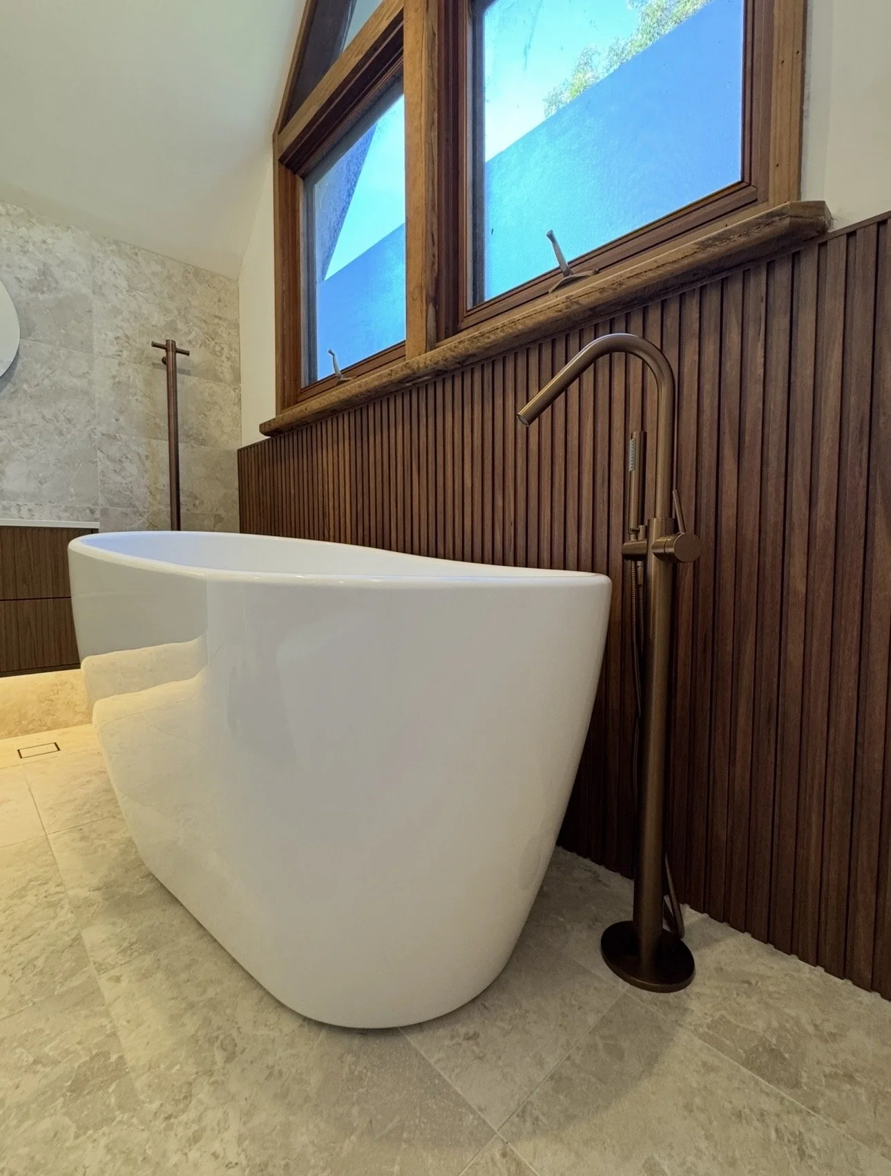 A modern bathroom featuring a white freestanding bathtub with a curved design, a bronze floor-mounted faucet, wooden wall paneling, large windows with a blue shade, and textured beige floor tiles.