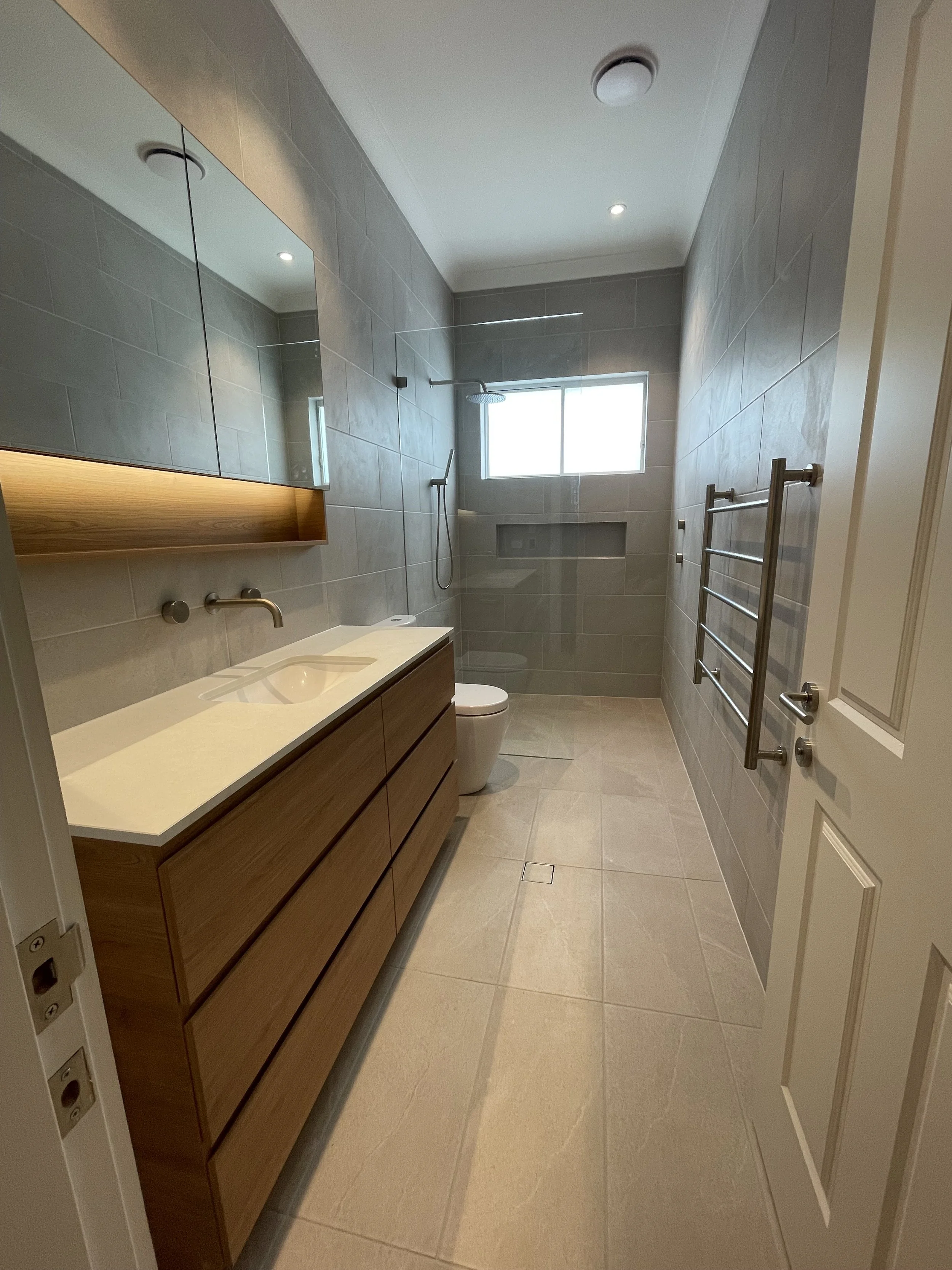 Modern bathroom with beige tile flooring, a wooden vanity with a white sink, large wall mirror, a window, and a walk-in shower with a rainfall showerhead.