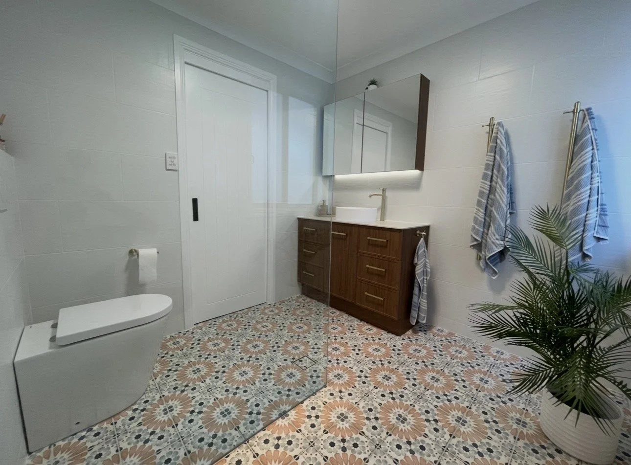Modern bathroom with white walls, a toilet, a wooden vanity with a white sink, a large mirror, and towels hanging on the wall. There is a potted plant in the corner and patterned floor tiles.