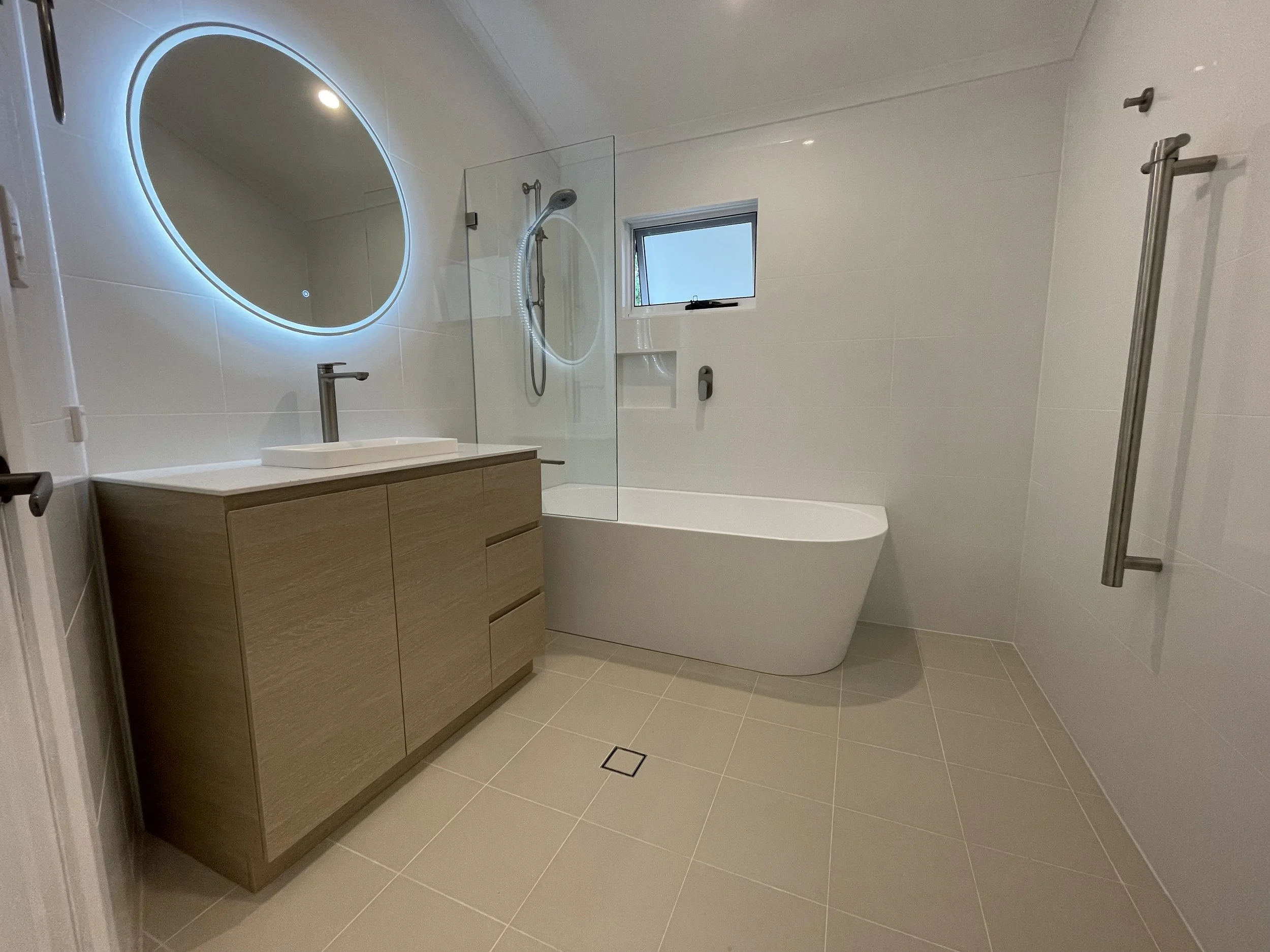Modern bathroom with a large oval mirror with LED lighting, a wooden vanity with a white sink, a walk-in shower with a handheld showerhead and glass partition, a small window, a bathtub, and beige tiled floor and walls.