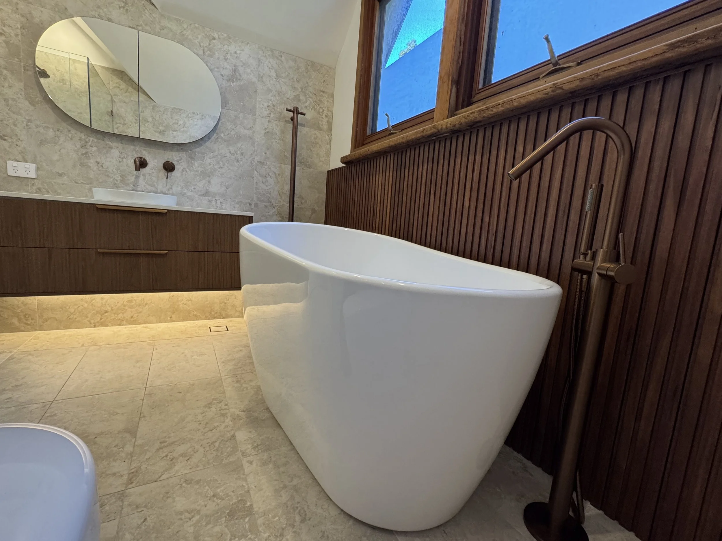 A bathroom features a white freestanding bathtub next to a wooden wall with vertical slats, a large window with wooden trim, a marble floor, a wooden vanity with a basin sink, an oval mirror, and modern bronze fixtures.