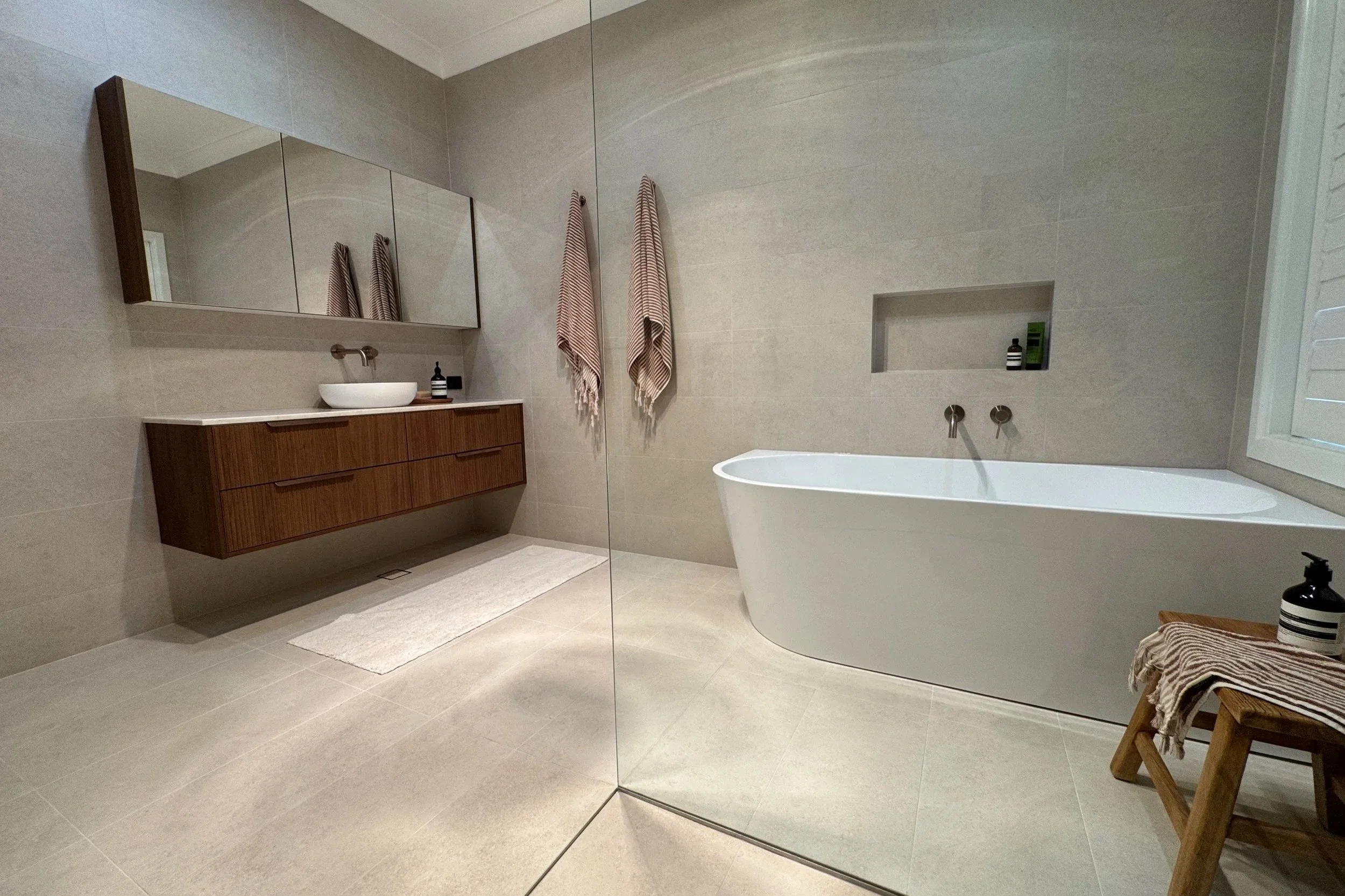 A modern bathroom with beige tiled walls and floor, a white freestanding bathtub, a wooden vanity with a vessel sink, large mirror, and hand towels hanging on the wall.