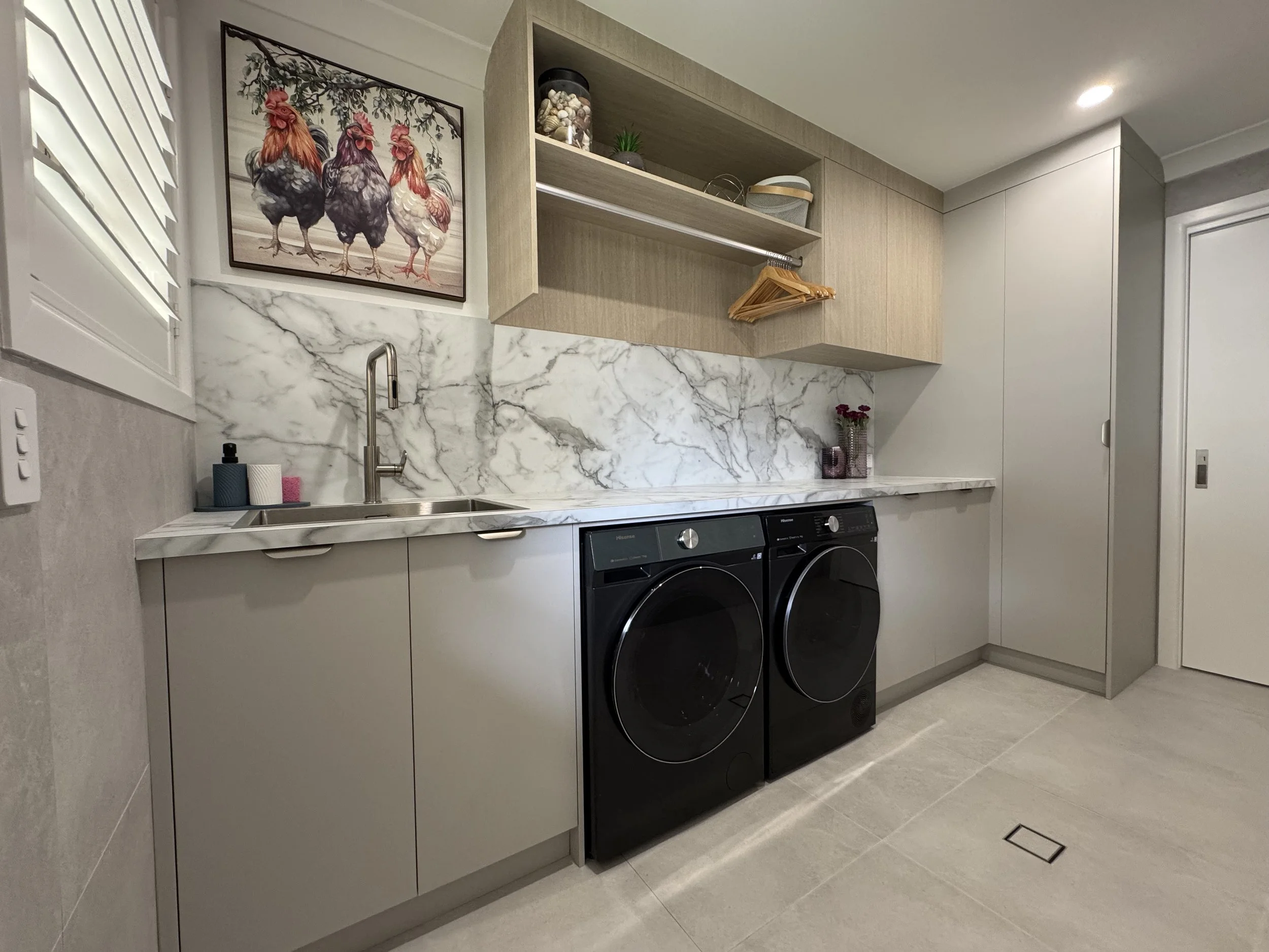 A modern laundry room with white cabinets, a marble countertop, and a marble backsplash. There is a stainless steel sink with a modern faucet, and a front-loading washer and dryer in black. Shelving above the countertop holds decorative items and sto
