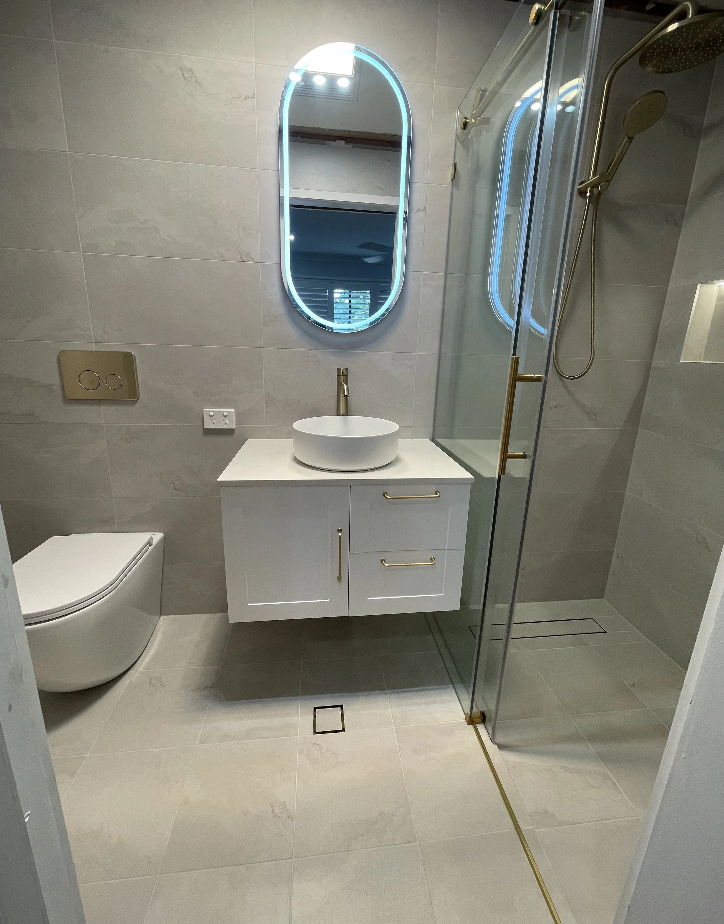 Modern bathroom with beige tiled walls and floor, a white vanity with a vessel sink, a backlit mirror, a glass shower enclosure with gold fixtures, and a wall-mounted toilet.