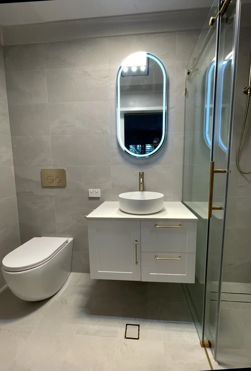 Modern bathroom with beige tiled walls and floor, a white toilet, a white vanity with a vessel sink and gold handles, an oval mirror with LED lighting, and a glass shower enclosure with gold fixtures.