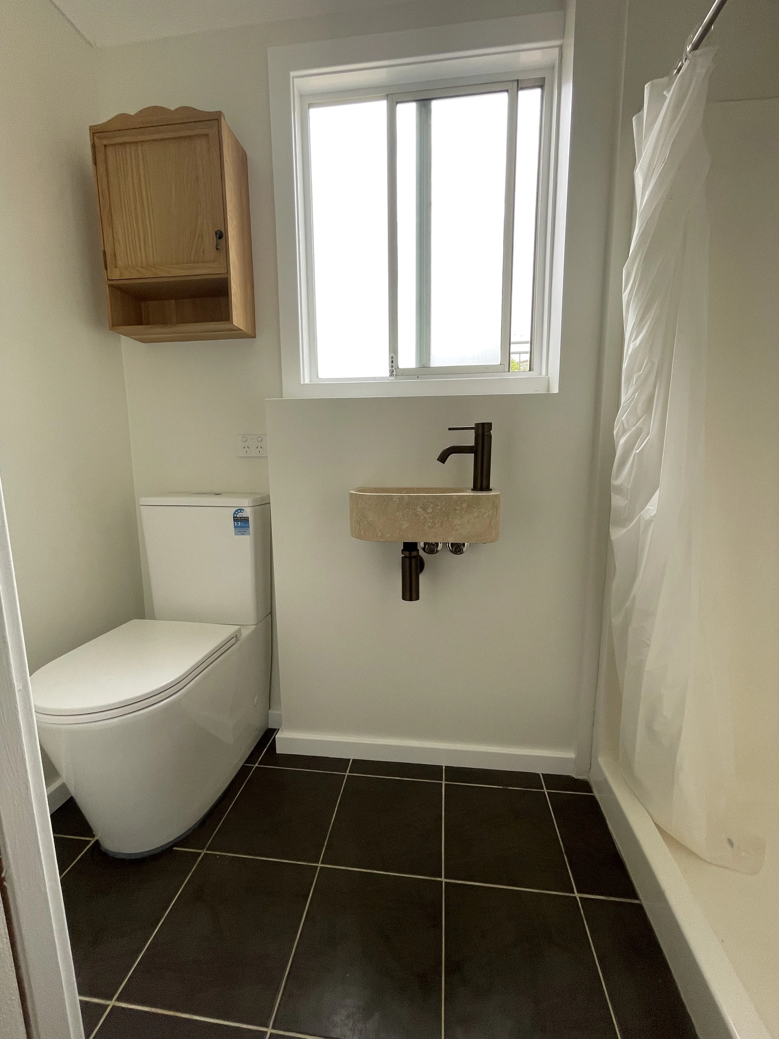 A small bathroom with a window, toilet, beige sink with a black faucet, black tiled floor, a wooden cabinet on the wall, and a white shower curtain.