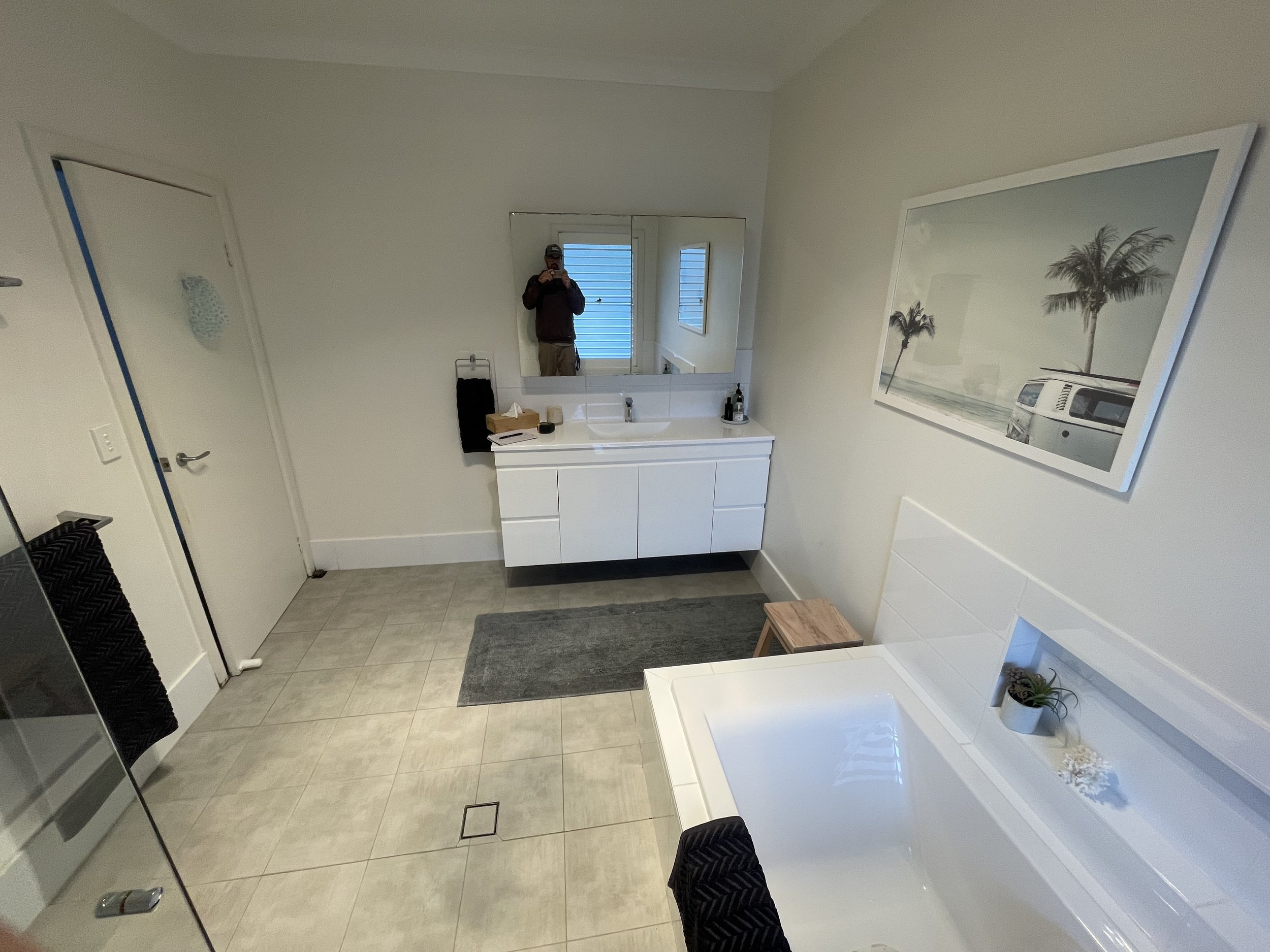 Bathroom with white vanity, large mirror, framed wall art, and a shower or bathtub with a white exterior and a small potted plant on it.