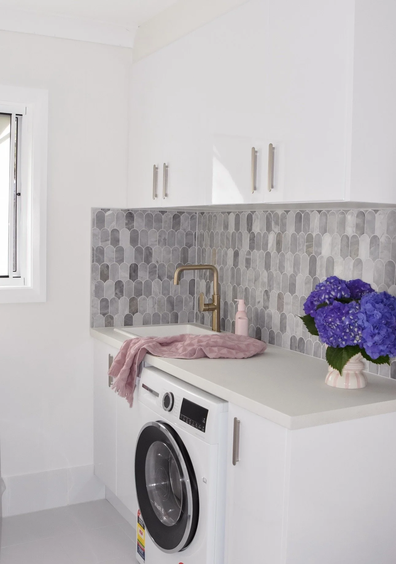 A laundry area with a front-loading washing machine, a white countertop, and decorative purple flowers in a pink vase, along with a pink cloth on the counter. The background features a white wall, gray patterned tiles, a gold faucet, and white cabine