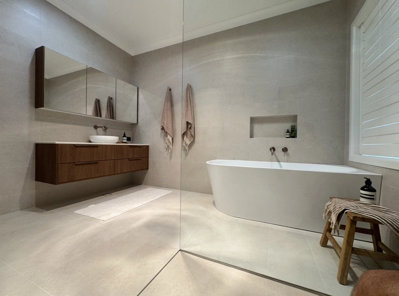 Modern minimalist bathroom with a white bathtub, wooden vanity with a vessel sink, large mirror, and neutral tiled walls and floor.