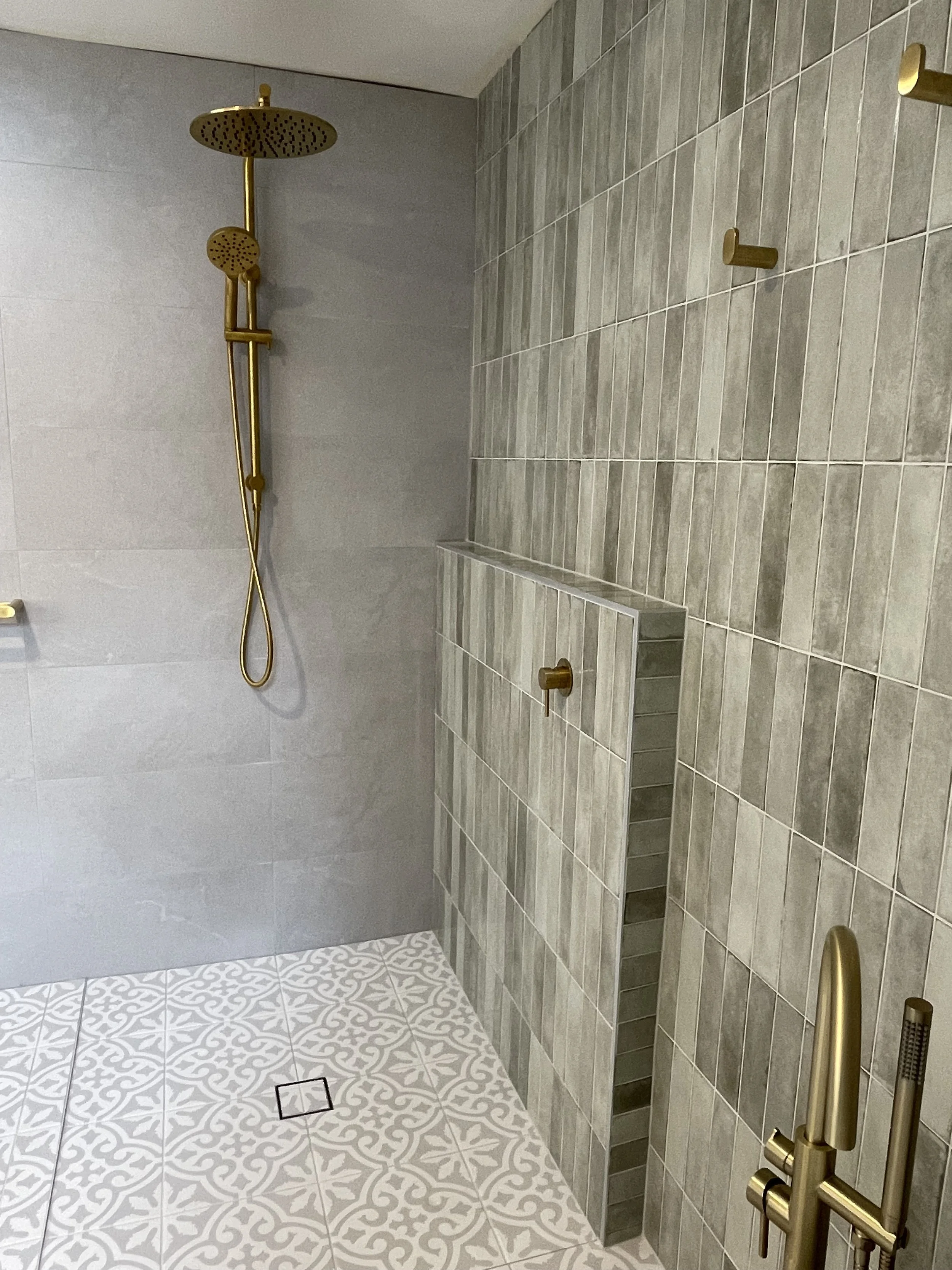 A modern bathroom shower area with gray tiles on the wall and patterned white and gray floor tiles. It features a gold showerhead and handheld shower fixture, gold wall hooks, and a built-in tiled bench with gray tiles.
