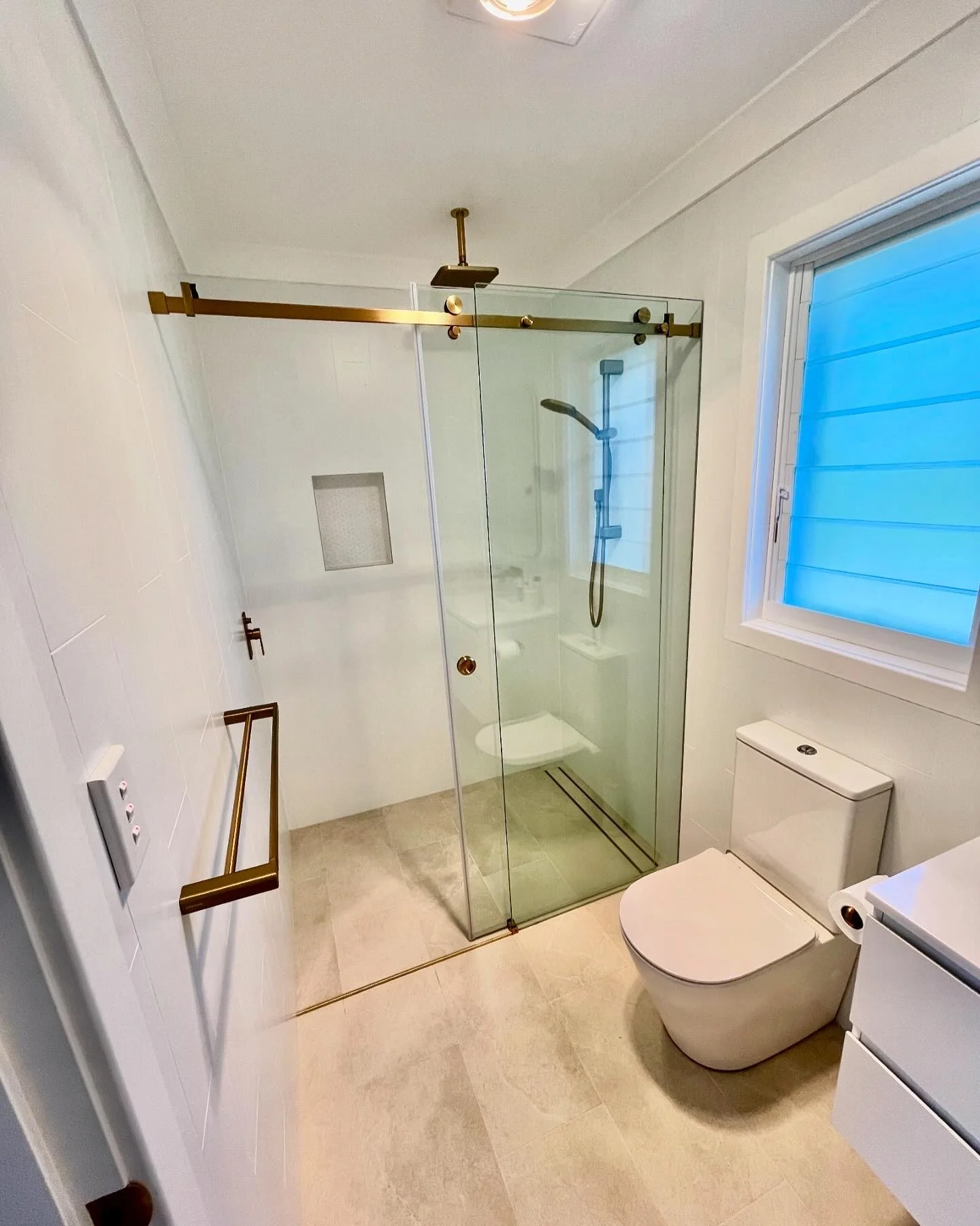 Modern bathroom with a glass shower enclosure, white toilet, window with blue blinds, and beige tiled floor.