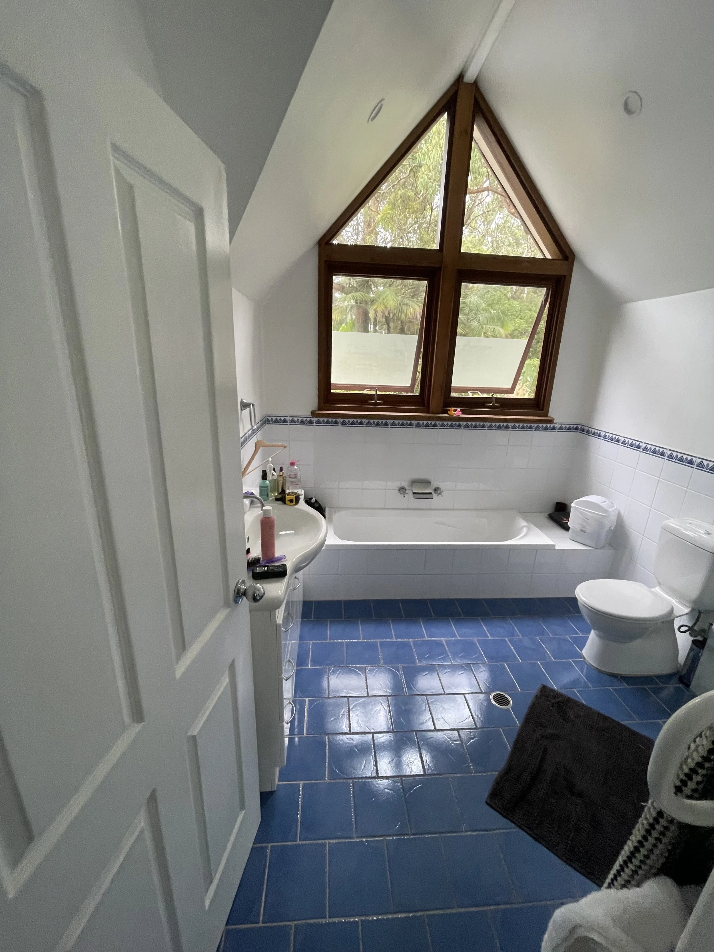 A bathroom with a sloped ceiling and a large triangular window, blue tiled floor, white walls with a decorative blue border, a white bathtub, a white toilet, and a sink with toiletries on it.