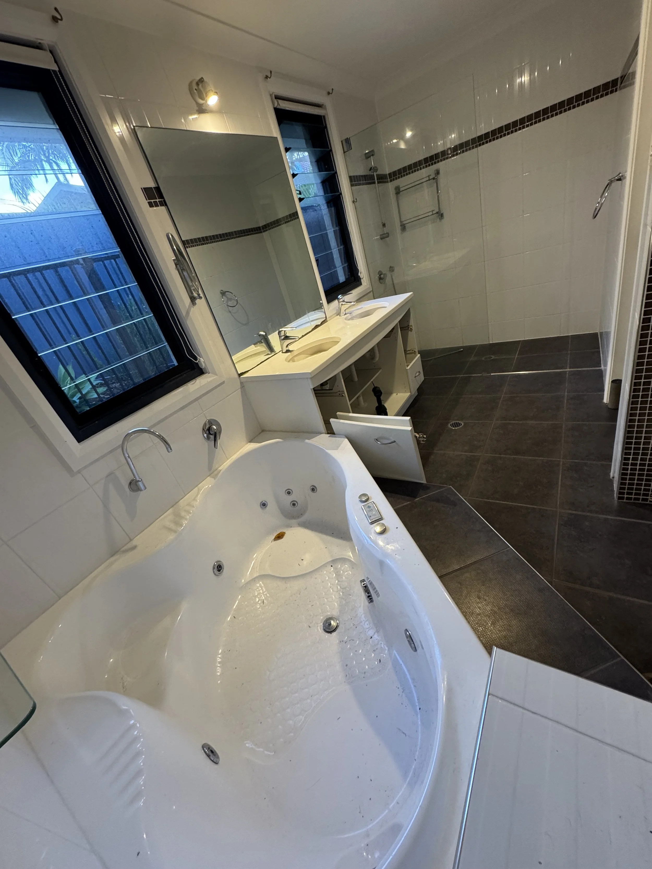 A spacious bathroom featuring a large white bathtub with multiple jets, a dual sink vanity with a large mirror, a walk-in shower with a glass enclosure, dark tiled flooring, and two windows providing natural light.