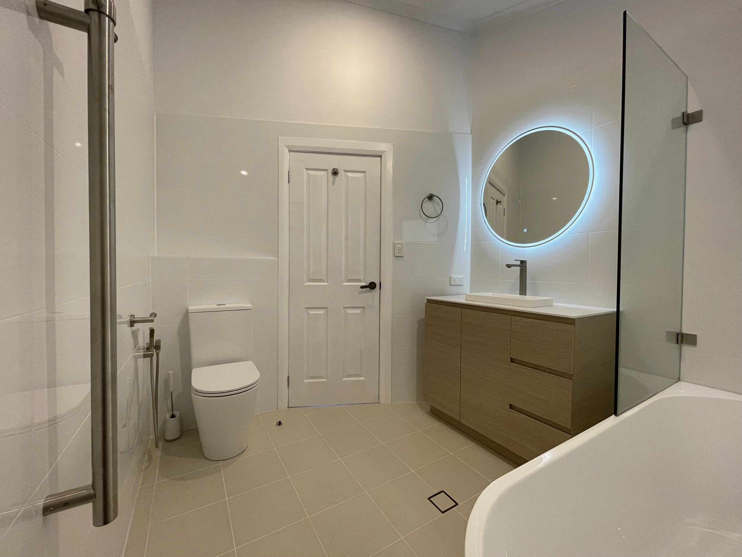 Modern bathroom with beige tiles, a white toilet, a wooden vanity with a white sink, a round illuminated mirror, a glass shower with a metal pole, and a small white bathtub.