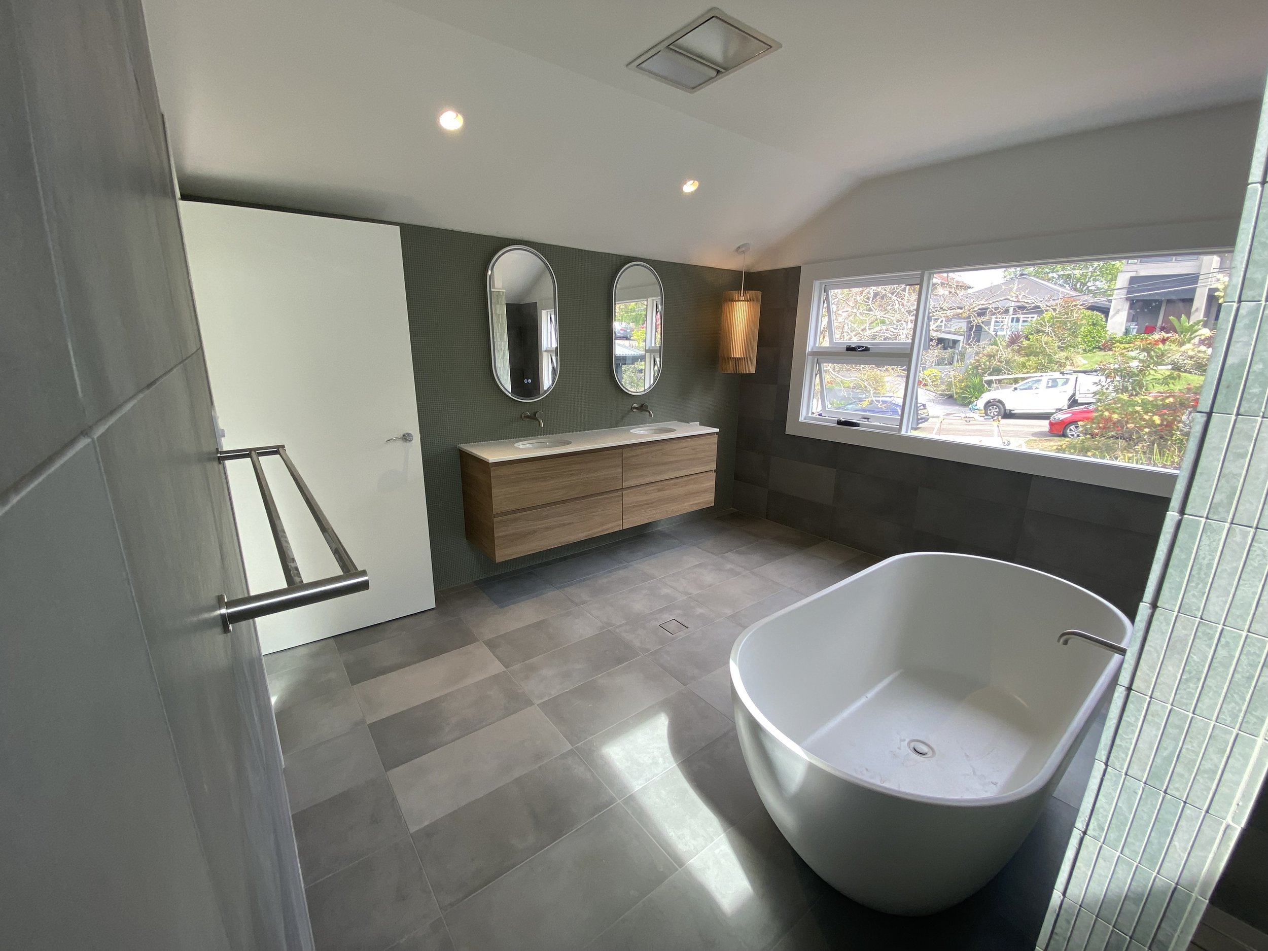 Modern residential bathroom with a freestanding bathtub, a wooden double vanity with two oval mirrors, a large window showing a neighborhood and green trees, tiled floors and walls, and ceiling lights.
