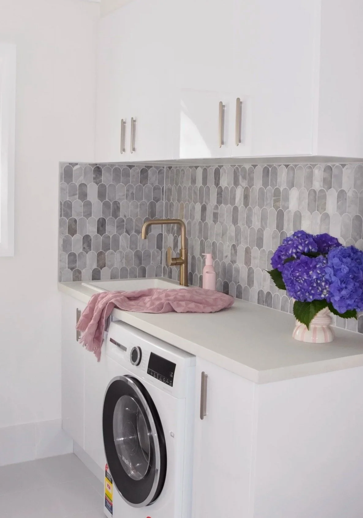 A laundry room with a white washing machine, a white countertop, a golden faucet, a pink soap dispenser, a pink cloth on the counter, and a vase with purple hydrangeas on the right. The background has a gray and white patterned tile backsplash and white cabinets.