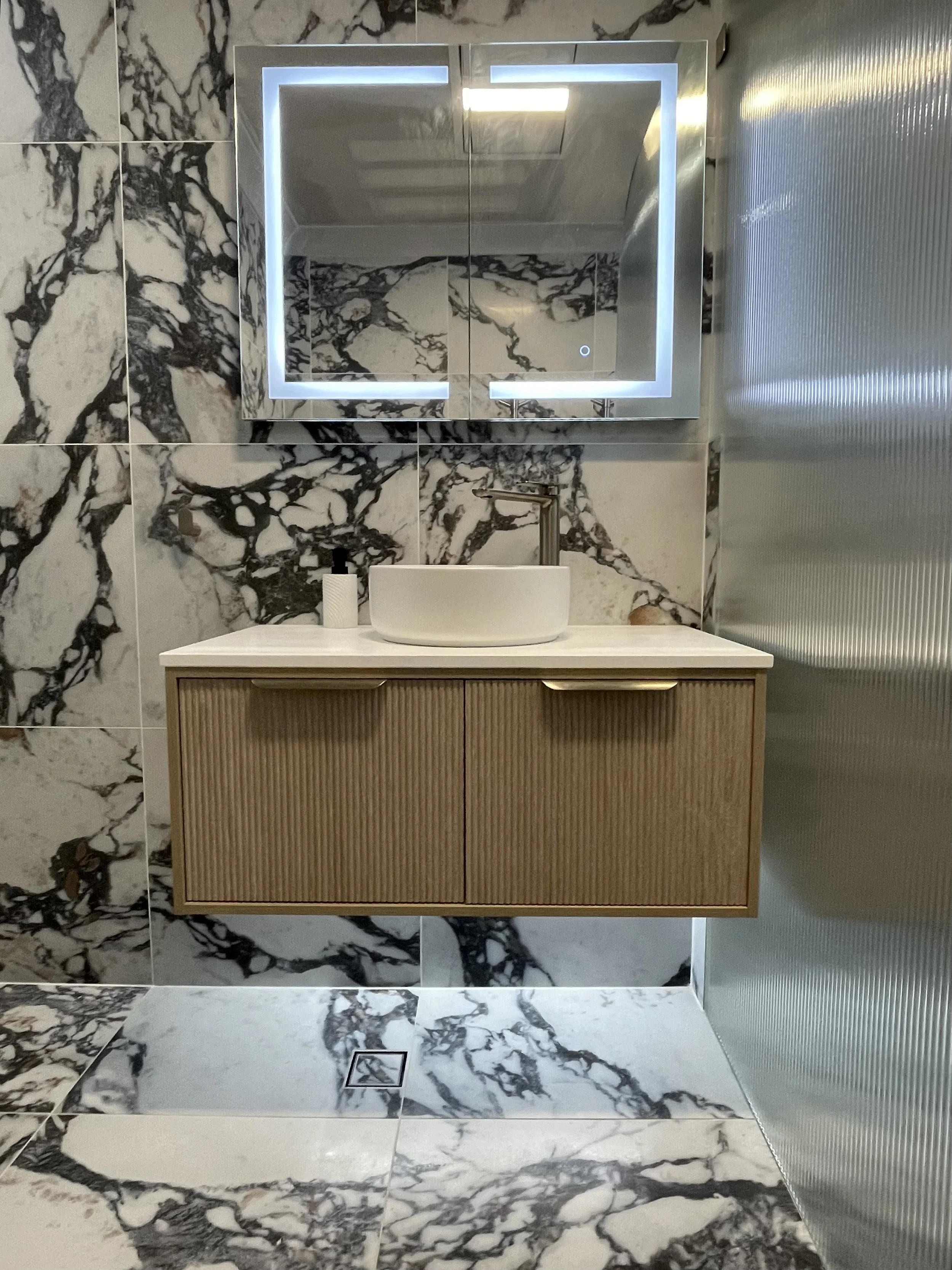 A marble wall with a mirror cabinet, a sink, and a soap dispenser in a modern bathroom with marble flooring.