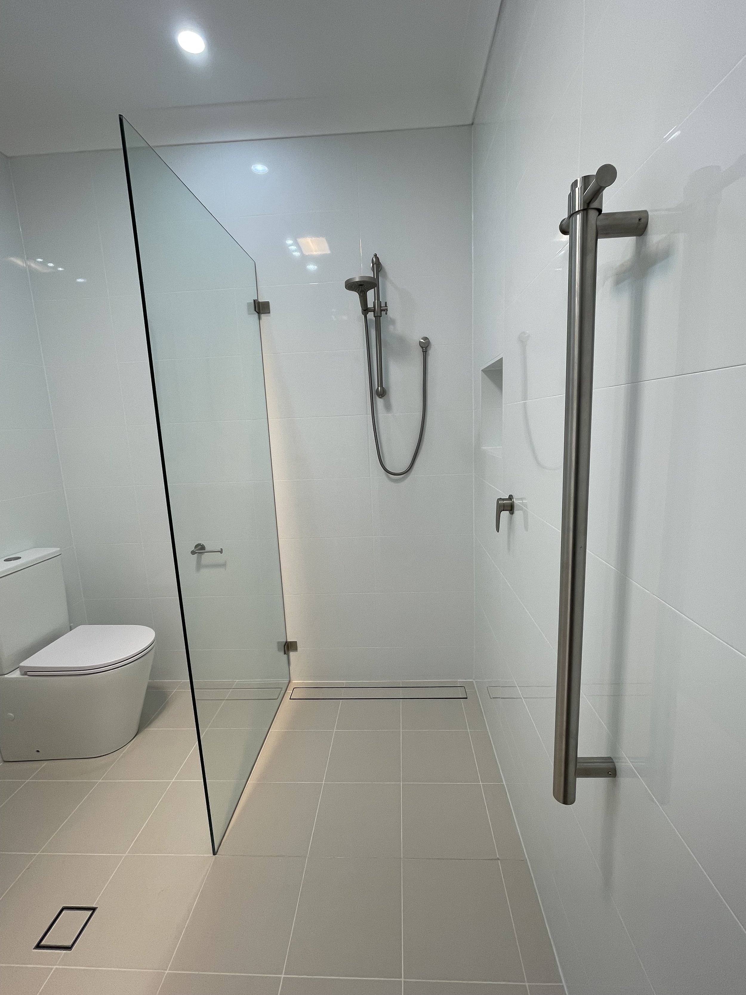 Modern bathroom with white tiled walls and floor, a toilet, a walk-in shower with a glass partition, a stainless steel grab bar, and ceiling lights.
