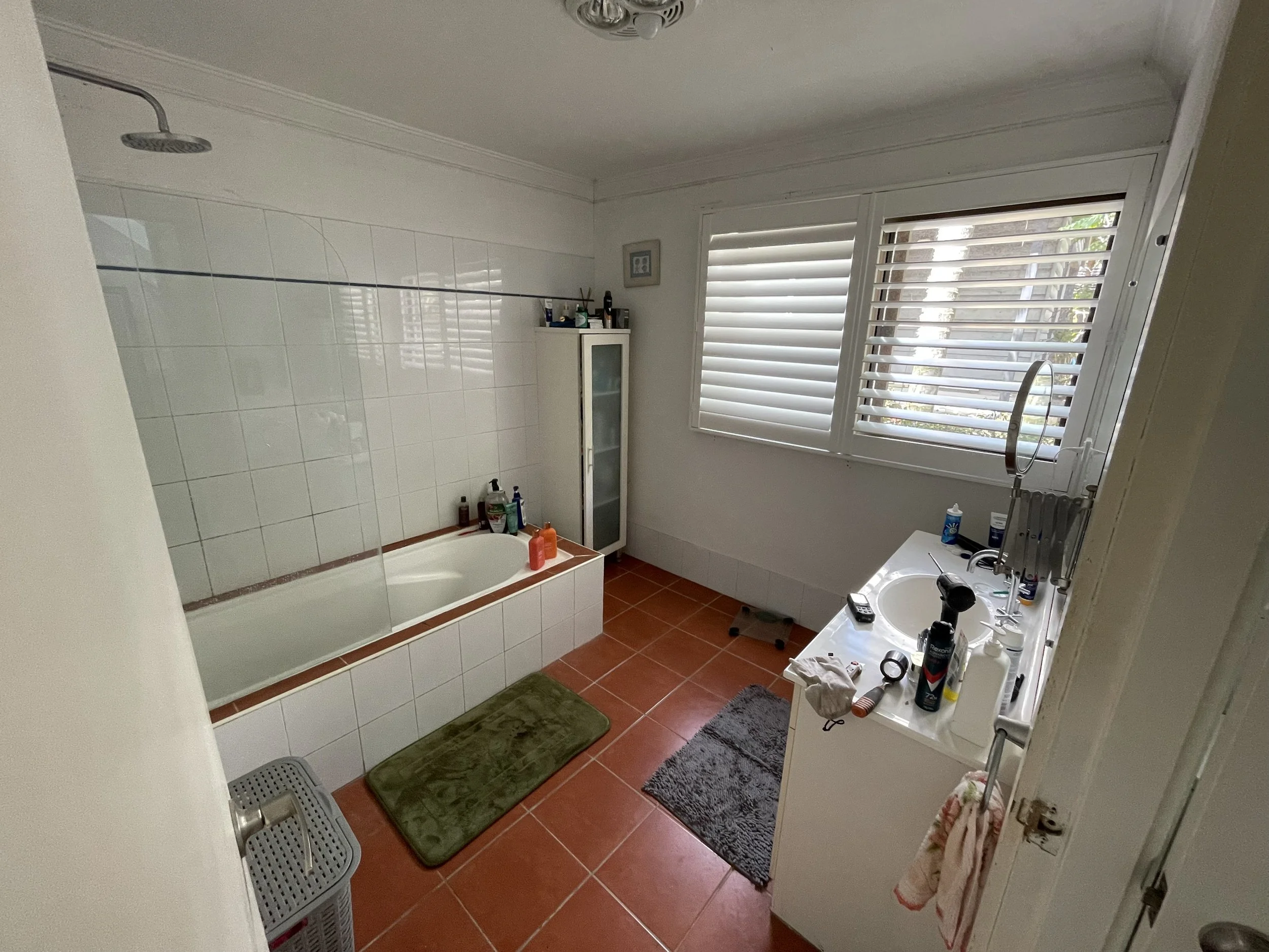 Bathroom with tile walls and floor, a bathtub, various toiletries, a sink with a faucet, a cabinet, and patterned bath mats, with sunlight coming through window shutters.
