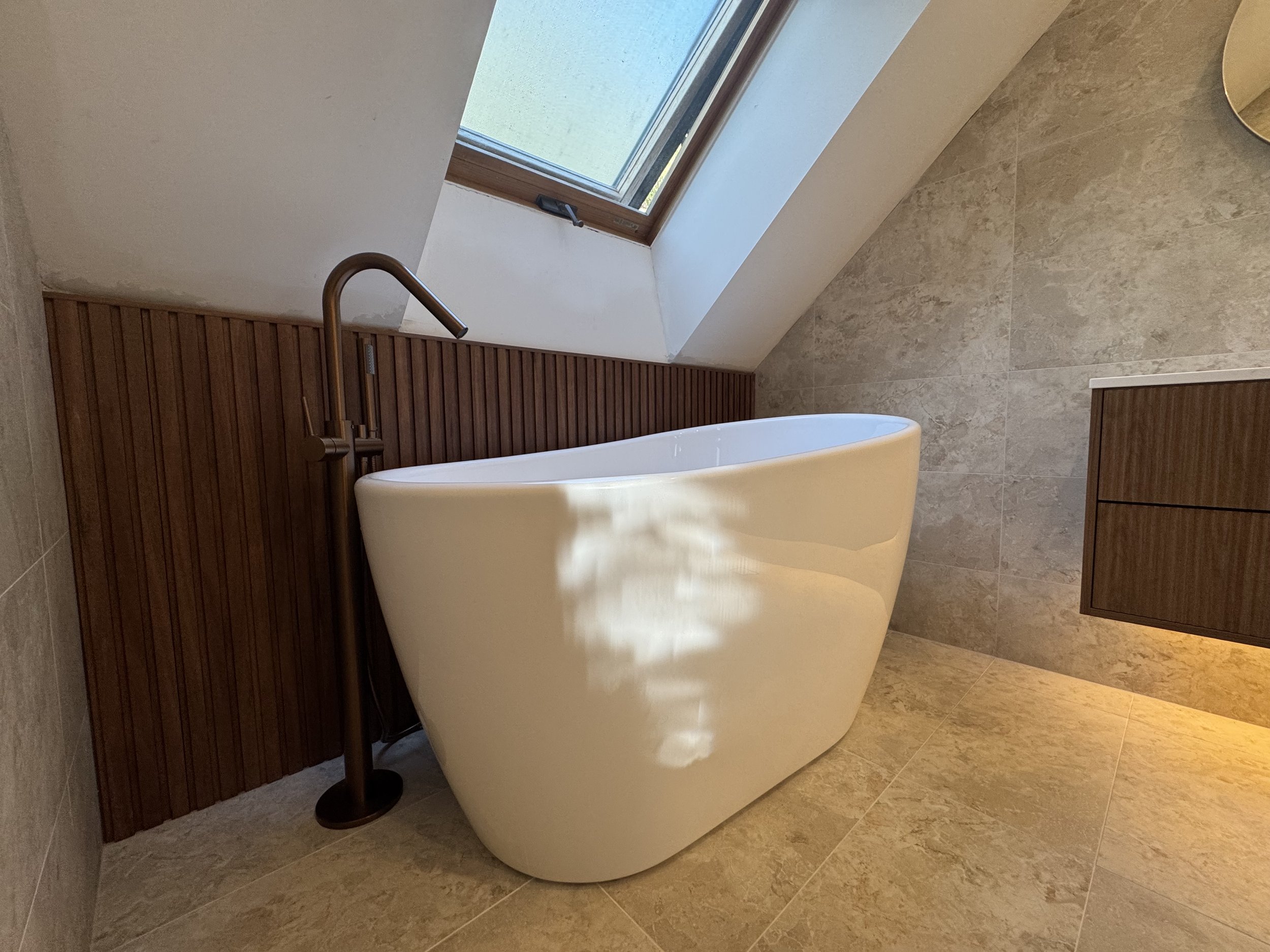 A bathroom with a white freestanding bathtub, a gold-colored wall-mounted faucet, beige tiled flooring, and a ceiling window letting in natural light.