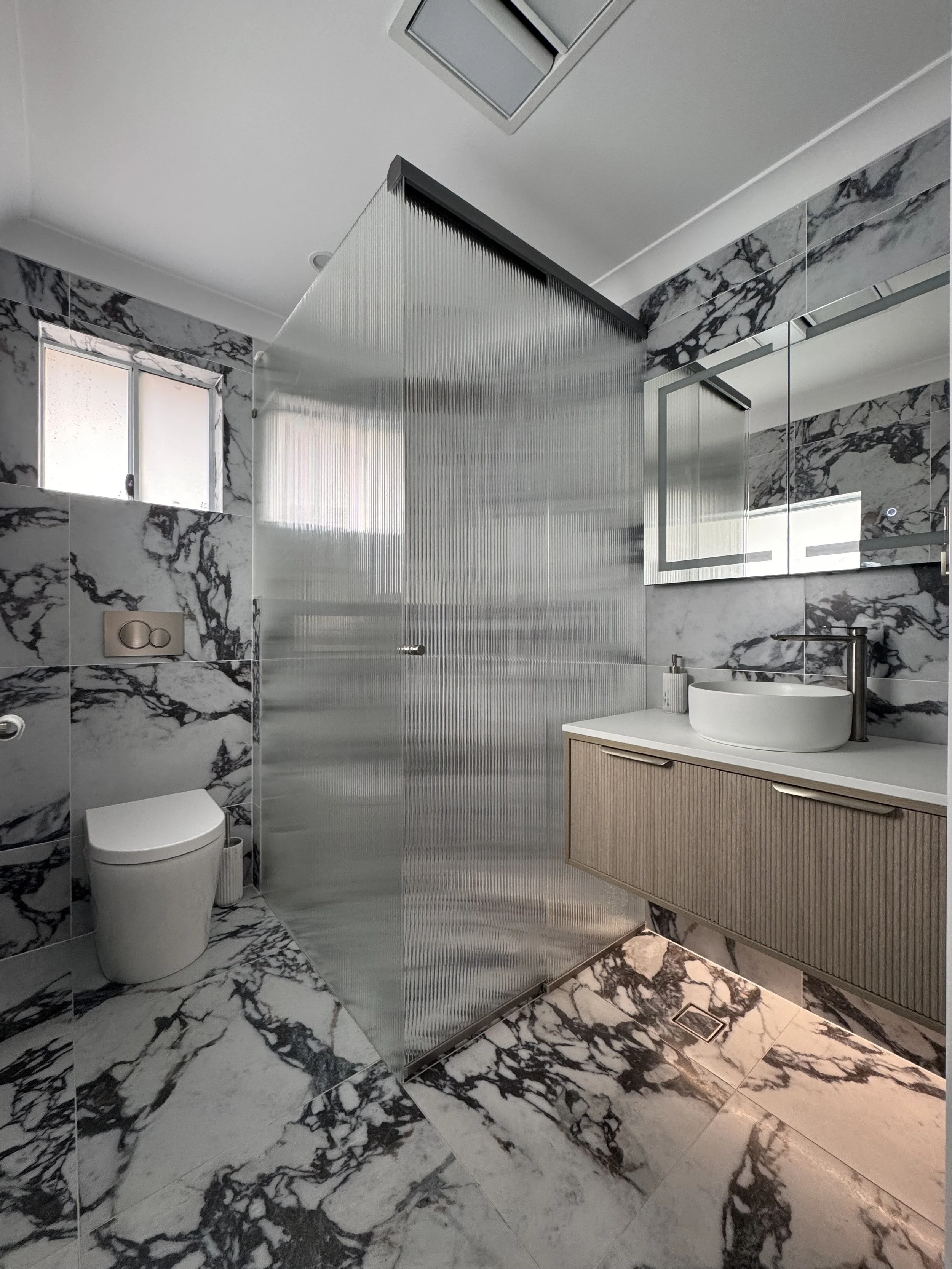 Modern bathroom with marble walls and flooring, a frosted glass shower enclosure, a wall-mounted toilet, and a vanity with a round vessel sink and a large mirror.