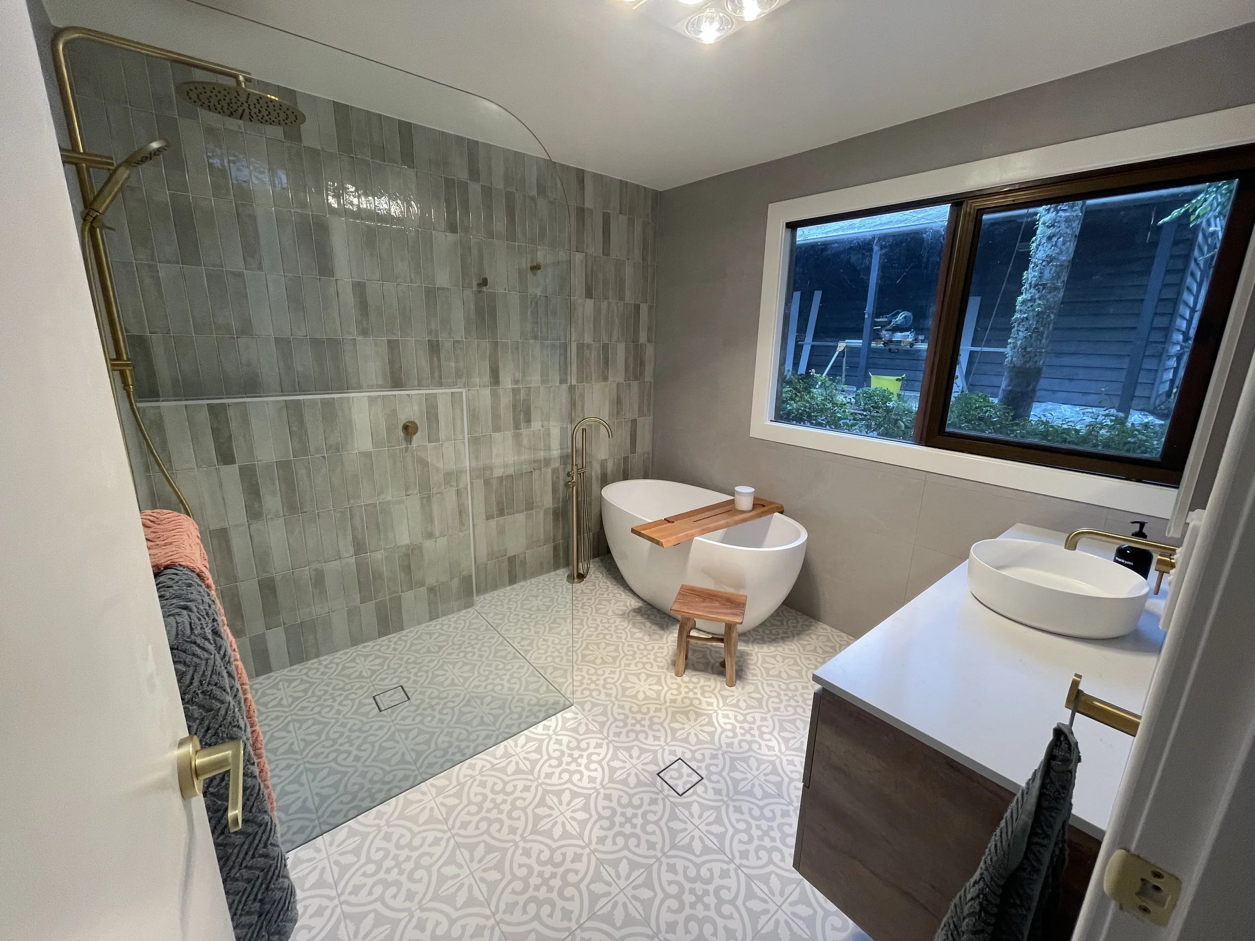 A modern bathroom with a walk-in shower with gray tiled walls and a clear glass door. The floor has patterned tiles, and there is a white freestanding bathtub with a wooden tray next to a large window showing outside greenery. The bathroom has a whit