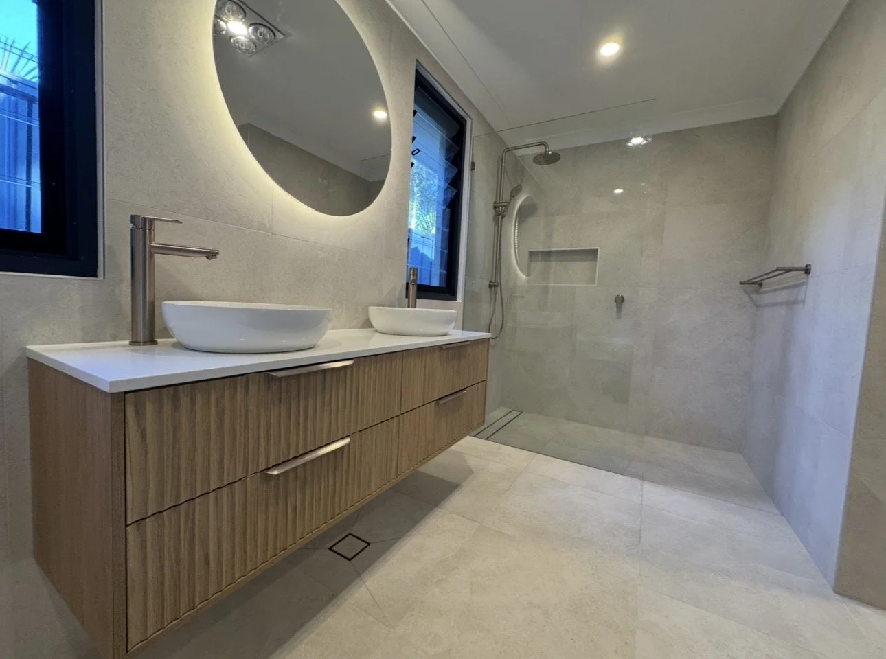Modern bathroom with double vessel sinks on a floating wooden vanity, large round backlit mirror, walk-in shower with a built-in shelf, and beige tiled walls and floor.