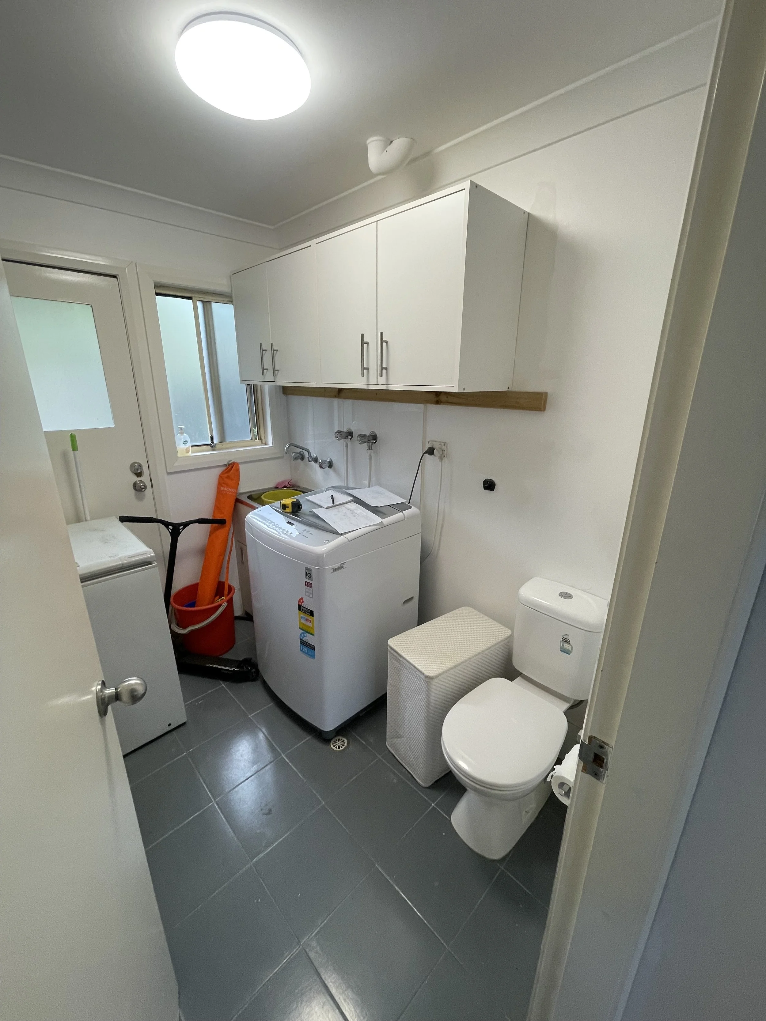 A small laundry room with a washing machine, a toilet, a white cabinet, and cleaning supplies.