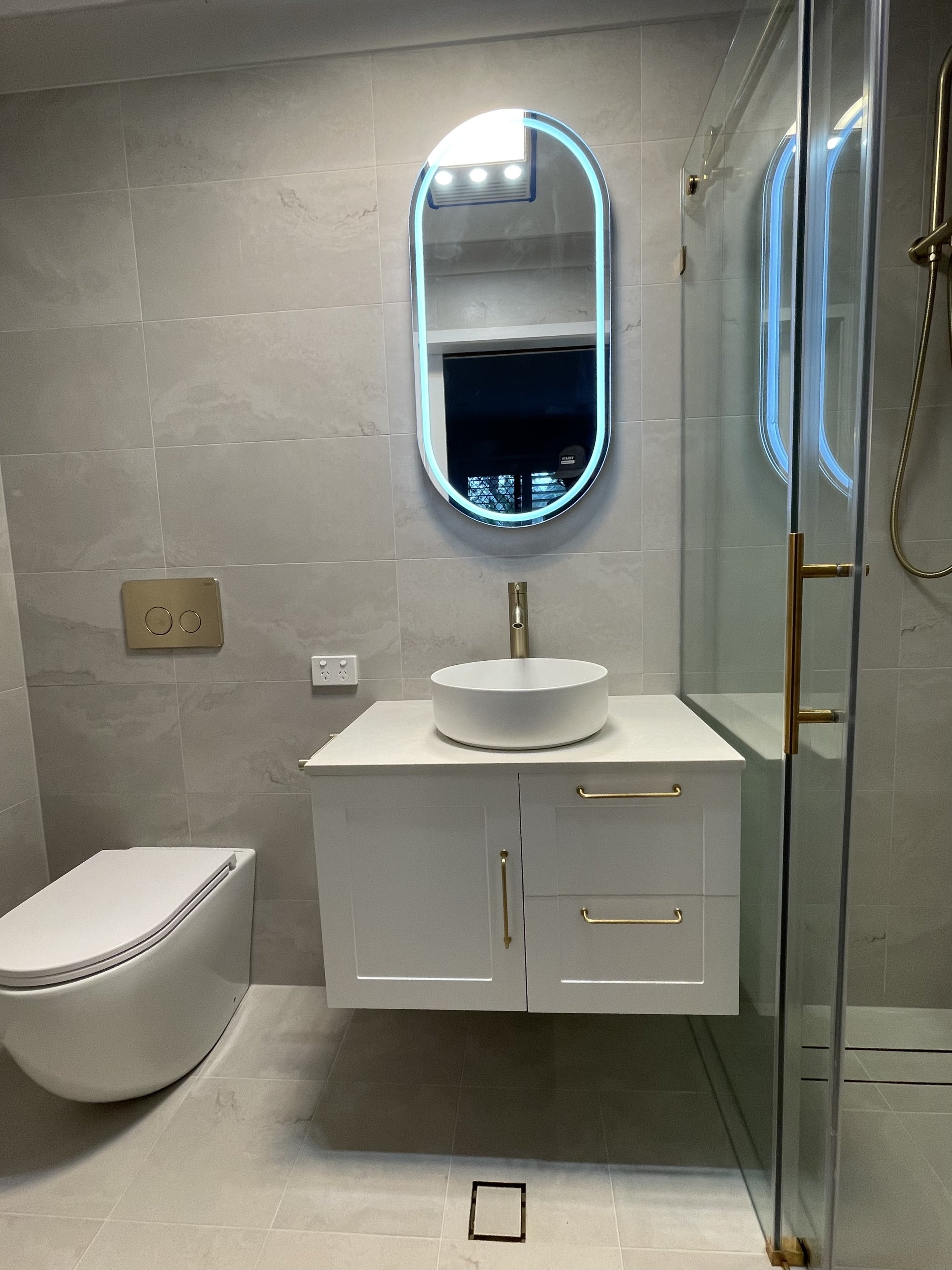 Modern bathroom with a wall-mounted oval mirror with LED lighting, a white vanity with a vessel sink and gold handles, a toilet, and a glass shower enclosure.