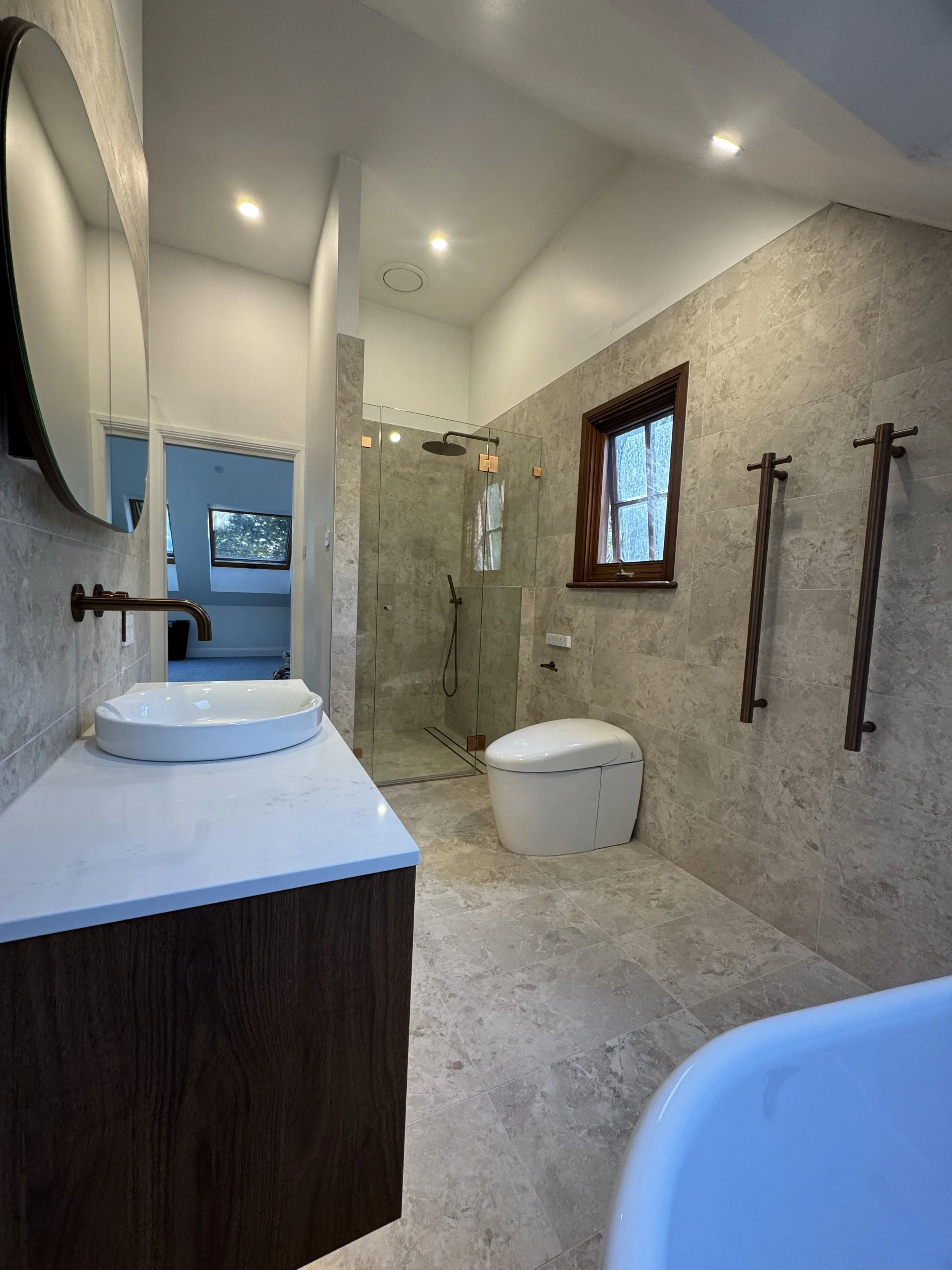 Modern bathroom with beige tile walls and floor, a walk-in shower with glass door and dark fixtures, a white toilet, a wooden-framed window, and a mirror reflecting part of the room.
