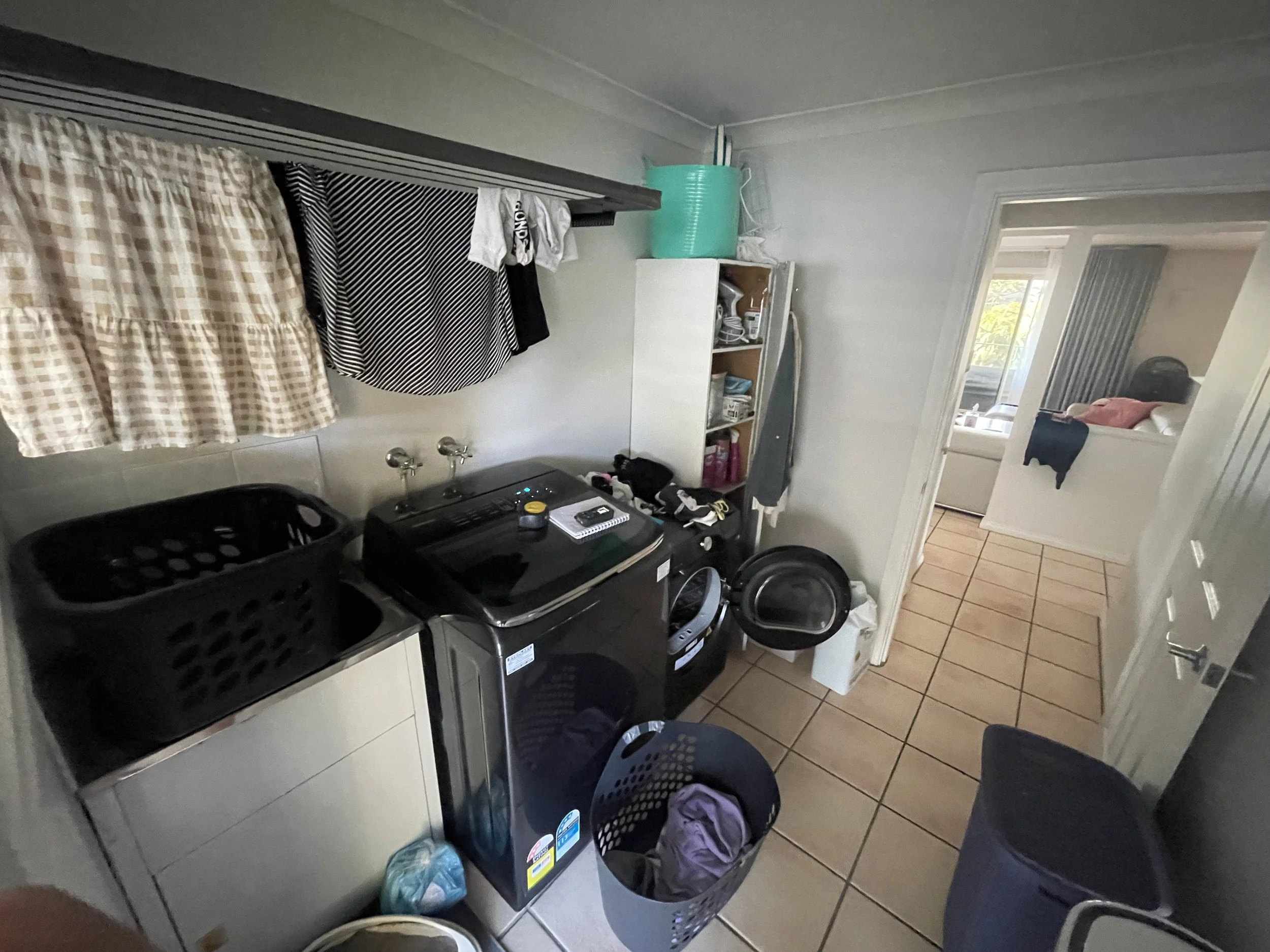 A laundry room with a washing machine, a dryer, laundry baskets, and laundry items. There are clothes hanging above and a mirror can be seen reflecting part of the room.