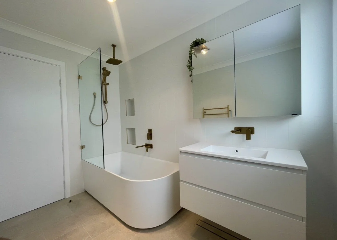 Modern bathroom with white bathtub, glass shower partition, wall-mounted fixtures, a large mirror above a white vanity, beige floor tiles, and a minimal design.