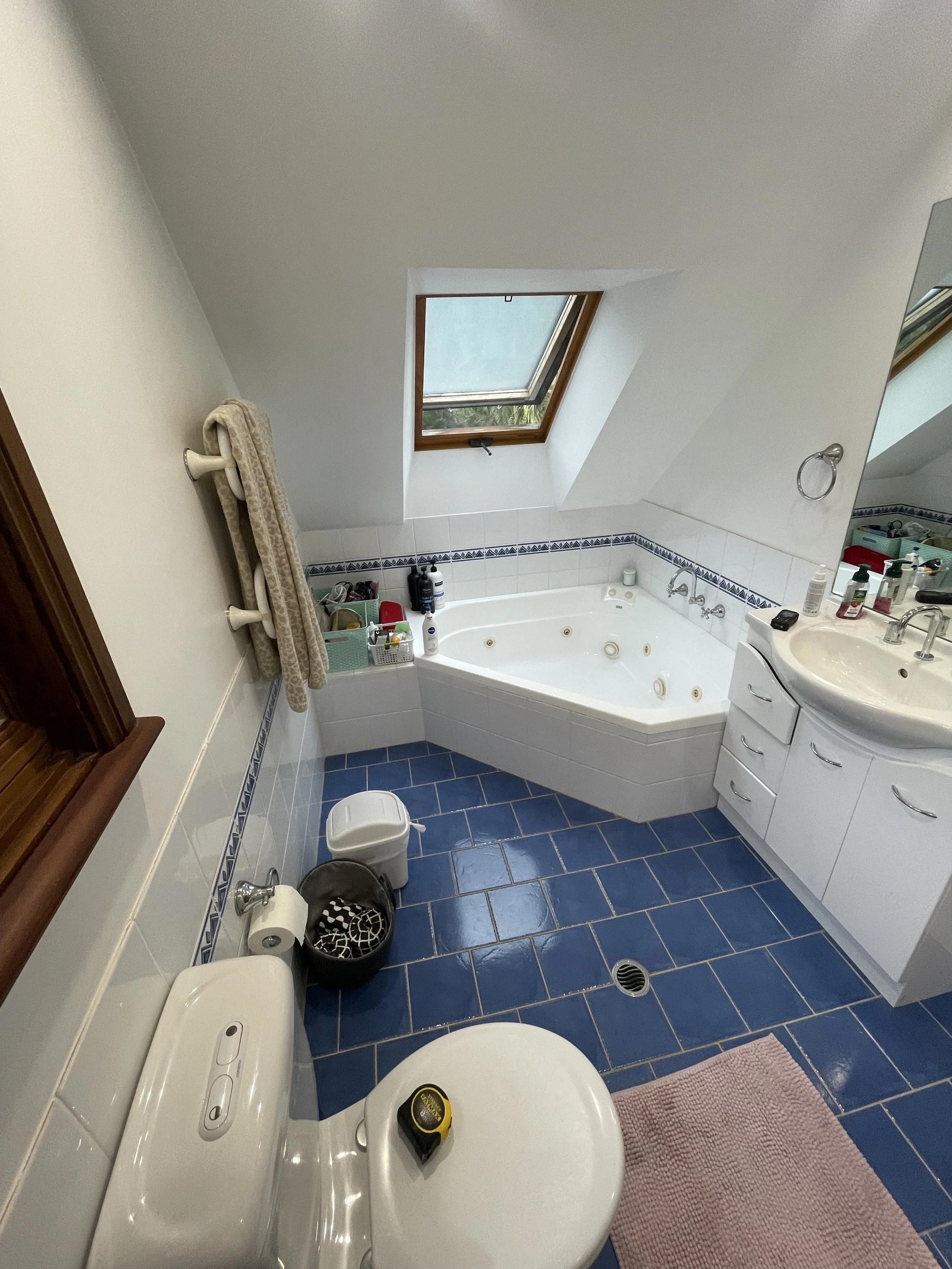 A small bathroom with a blue tiled floor, a white corner bathtub with jet nozzles, a skylight window in the ceiling, a white vanity with toiletries, a towel rack with towels, a toilet with a toilet paper roll, and a pink bath mat.