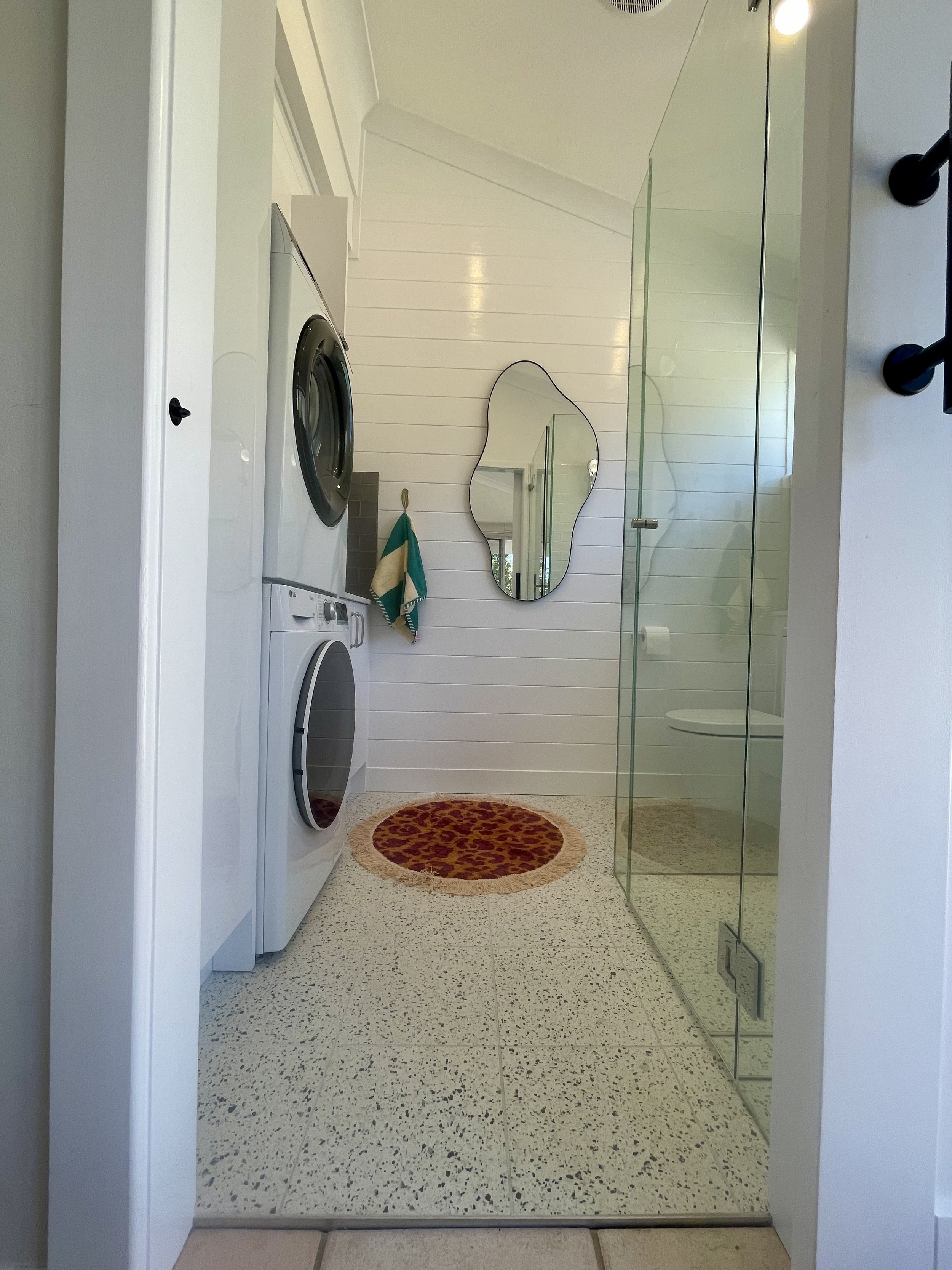 A laundry room with a washing machine and dryer stacked on top of each other, a mirror on the wall, a towel hanging, a small rug, and a glass door leading to another room.