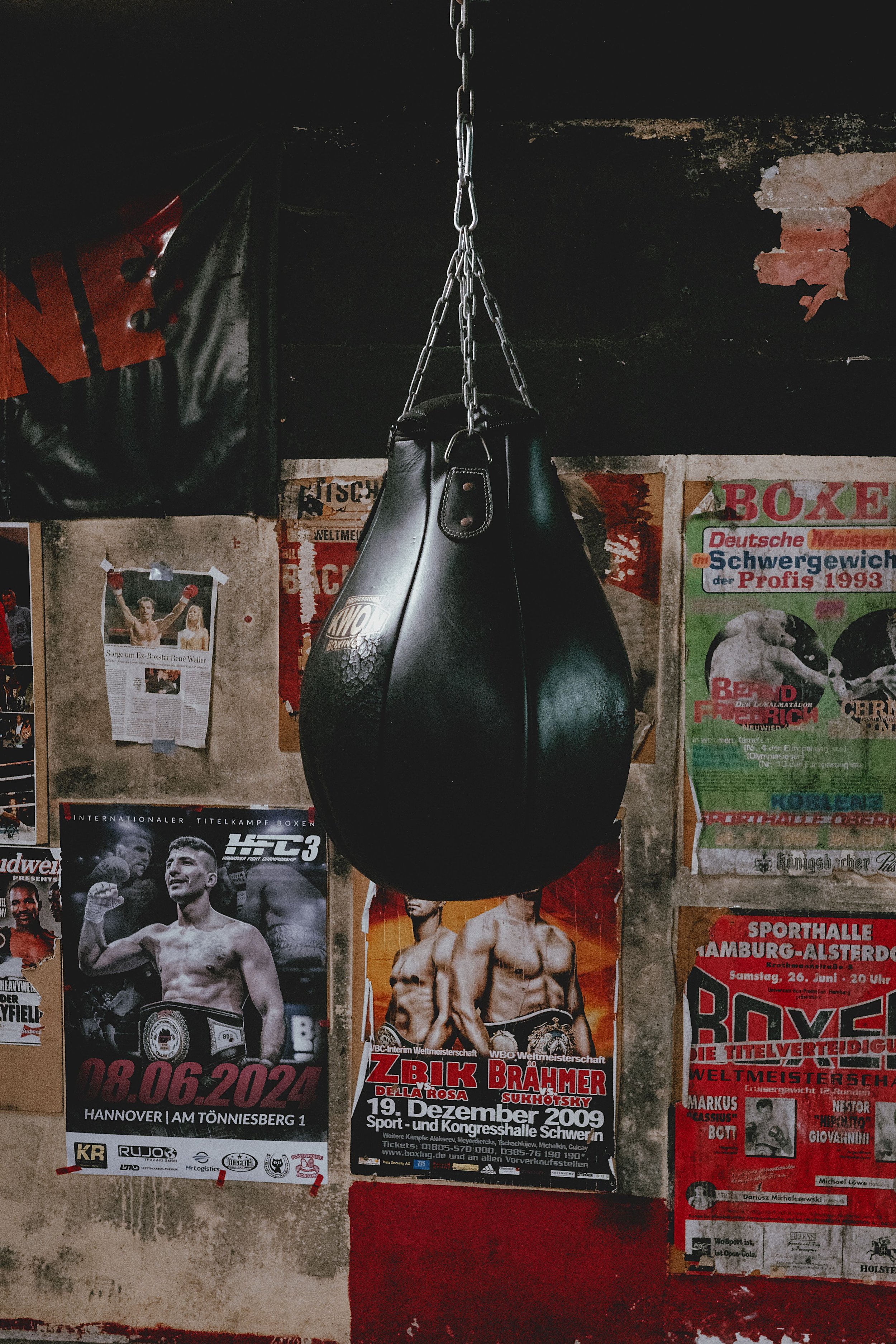 A black hanging punching bag mounted on a chain in a gym, with various posters on an old wall behind it, including boxing event posters and a boxer celebrating.