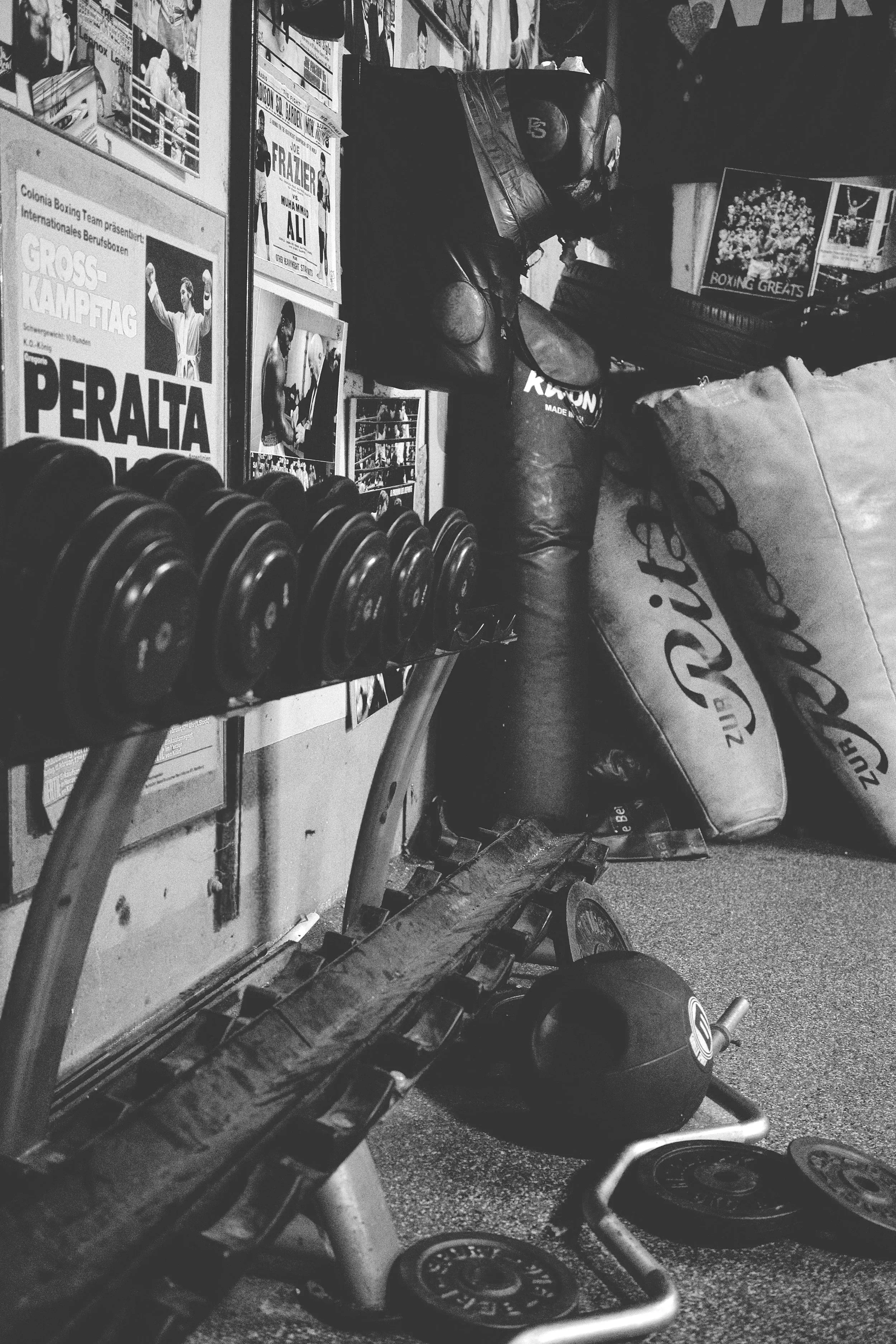A boxing gym corner with boxing gloves on a stand, a punching bag, and various posters on the wall, including promotional boxing posters and a framed photo, with a rack of dumbbells in the foreground.